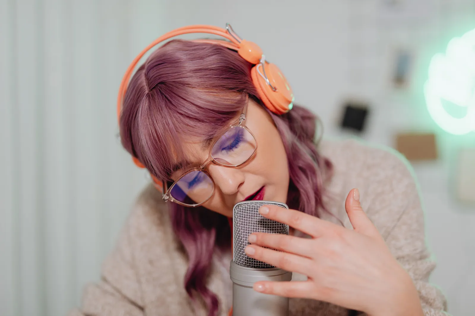 A young woman with vibrant hair and glasses recording audio with a microphone indoors