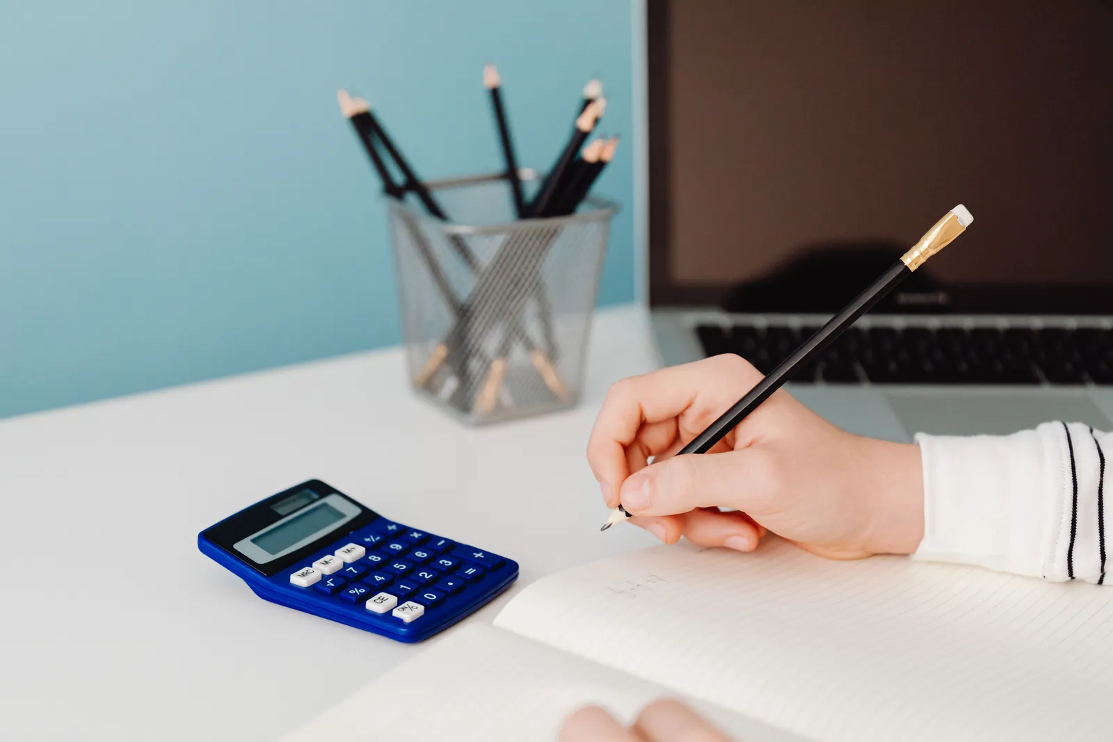 Close-up of a hand writing in a notebook beside a calculator and laptop on a desk