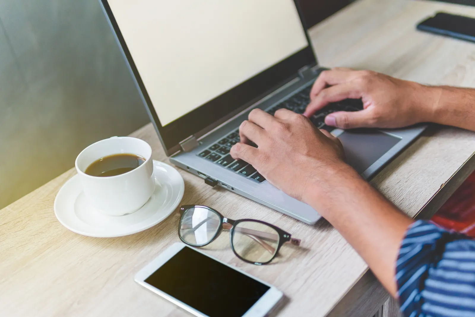 A man is typing on a laptop at a desk with coffee, phone, and glasses, illustrating a remote work setup