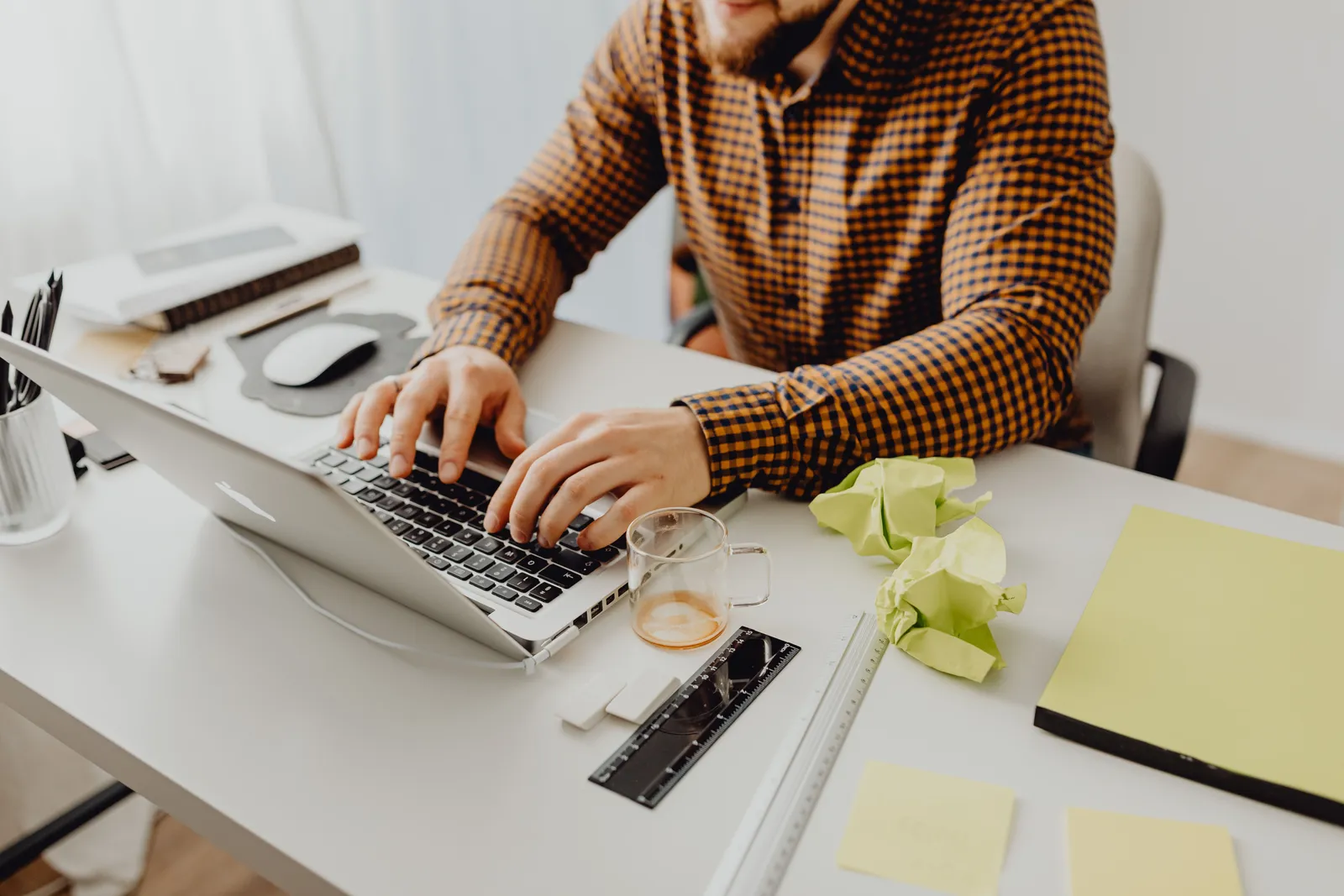 A man wearing a checkered shirt types on a laptop at his office desk
