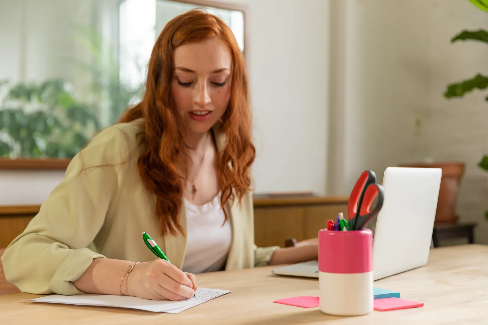 Woman with red hair writing on paper at home office desk with a laptop