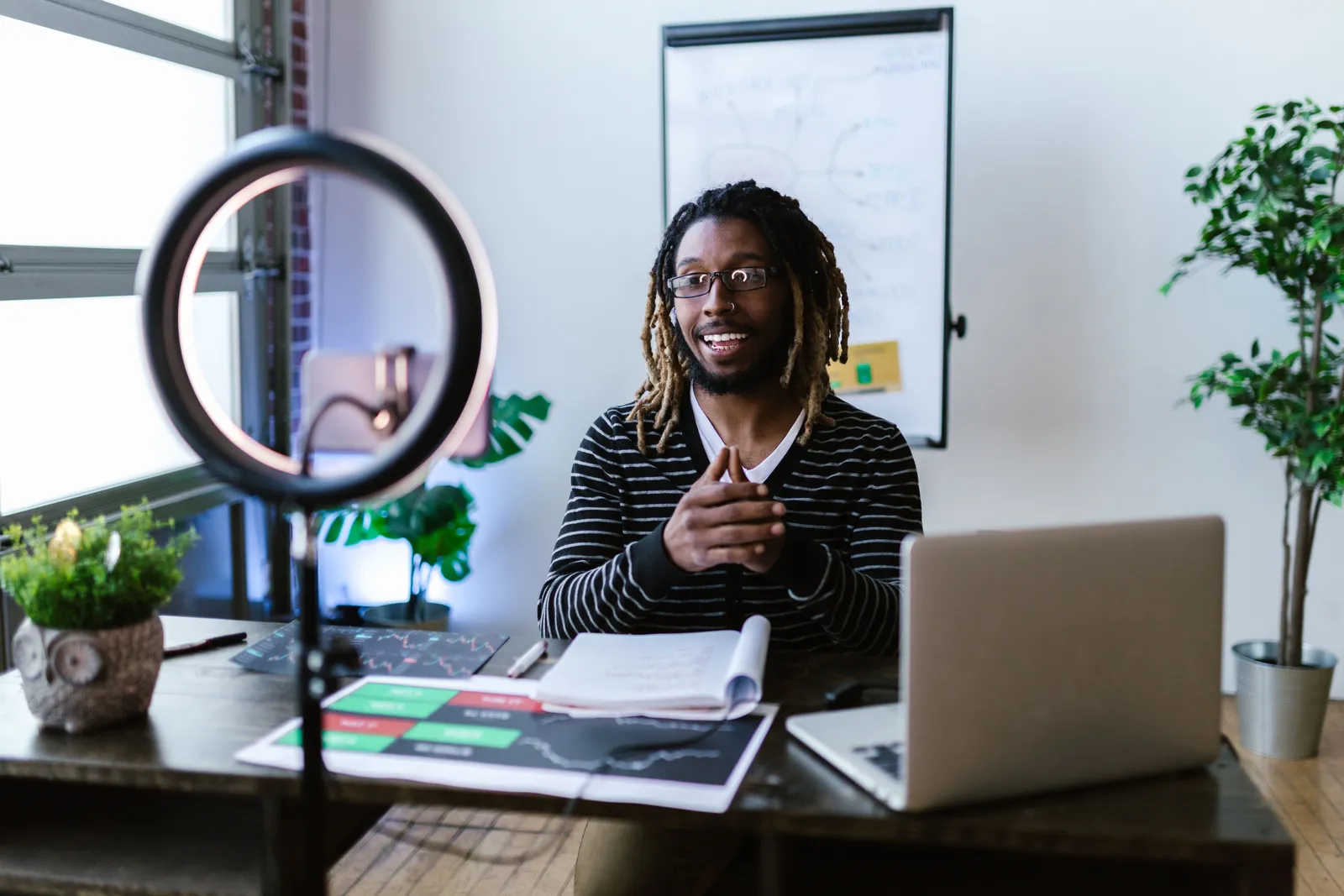 Young man with dreadlocks creating a video blog using smartphone and ring light in home office