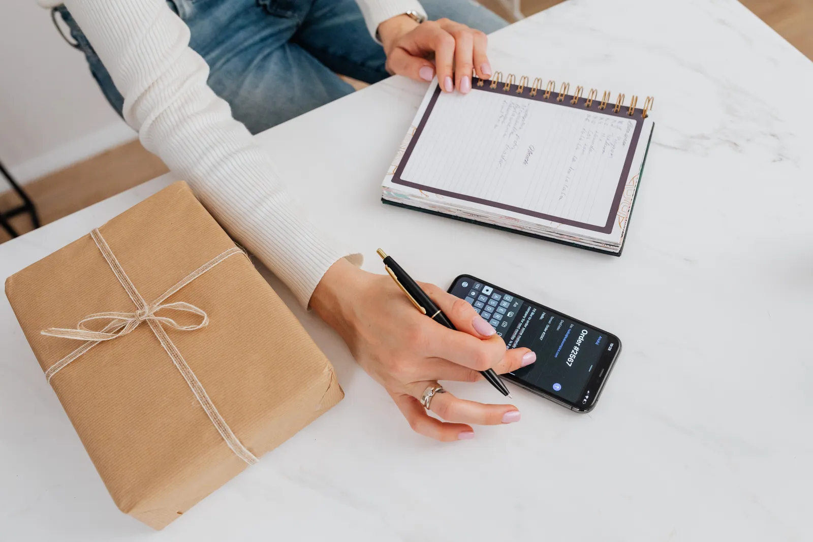 Person writing notes in a notebook beside a smartphone on a desk with a wrapped gift