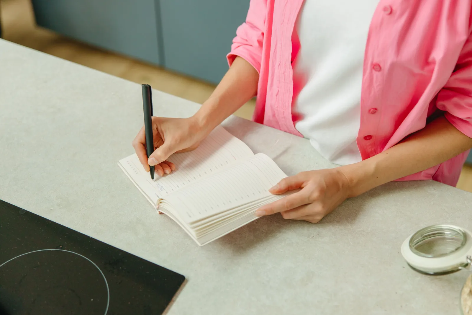 A woman in a pink shirt writing in a notebook on a kitchen counter with a pen
