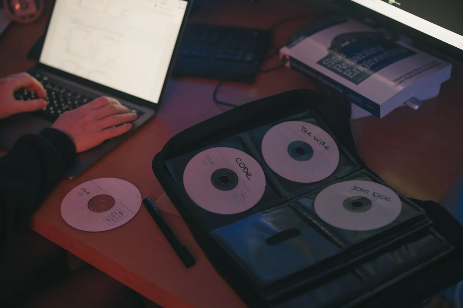 A person works on a laptop beside a set of labeled CDs on a desk