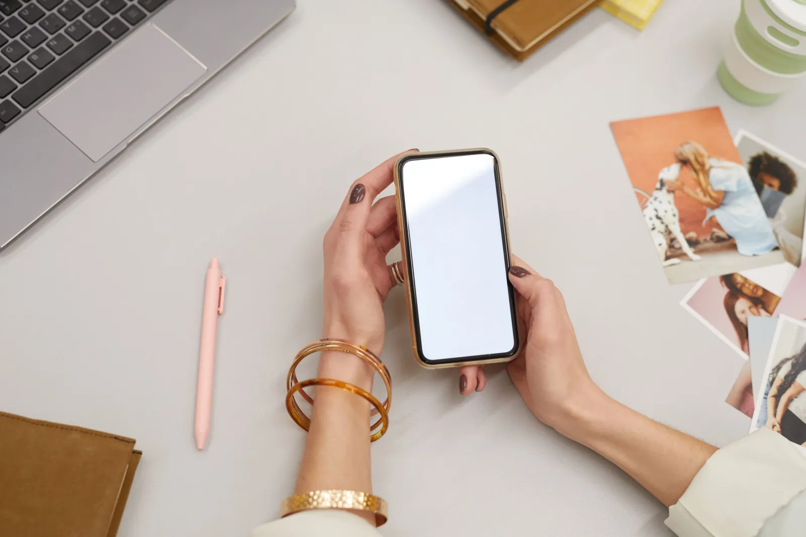 Close-up of a woman holding a smartphone on a desk with a laptop and photos, capturing modern workspace vibes