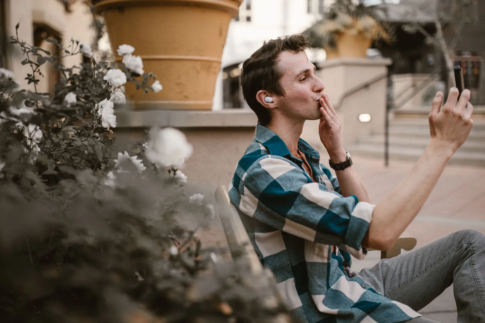 A man sitting in a park, making a video call on his phone, wearing earbuds