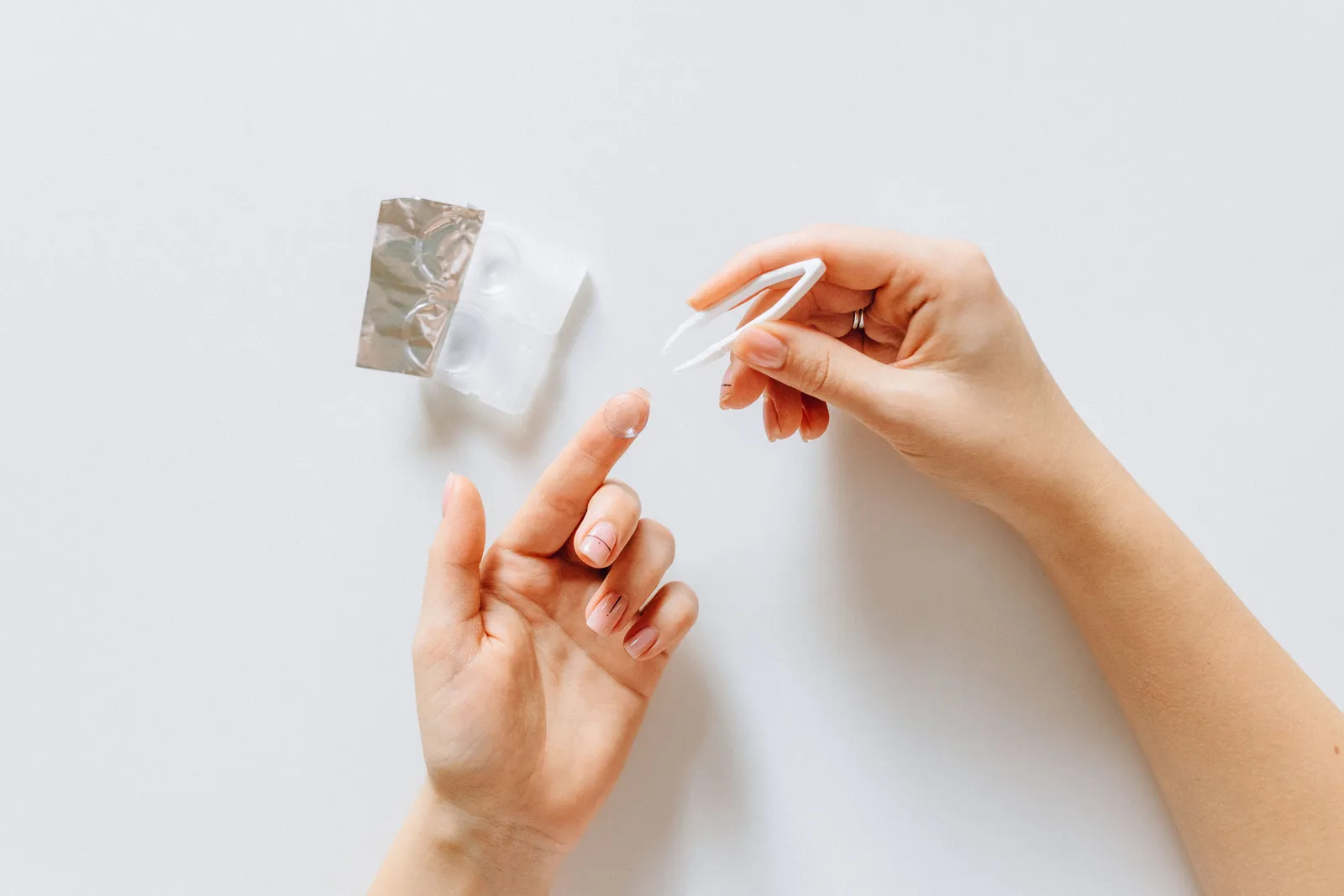 Hands holding a contact lens with tweezers next to a contact lens case on a white background