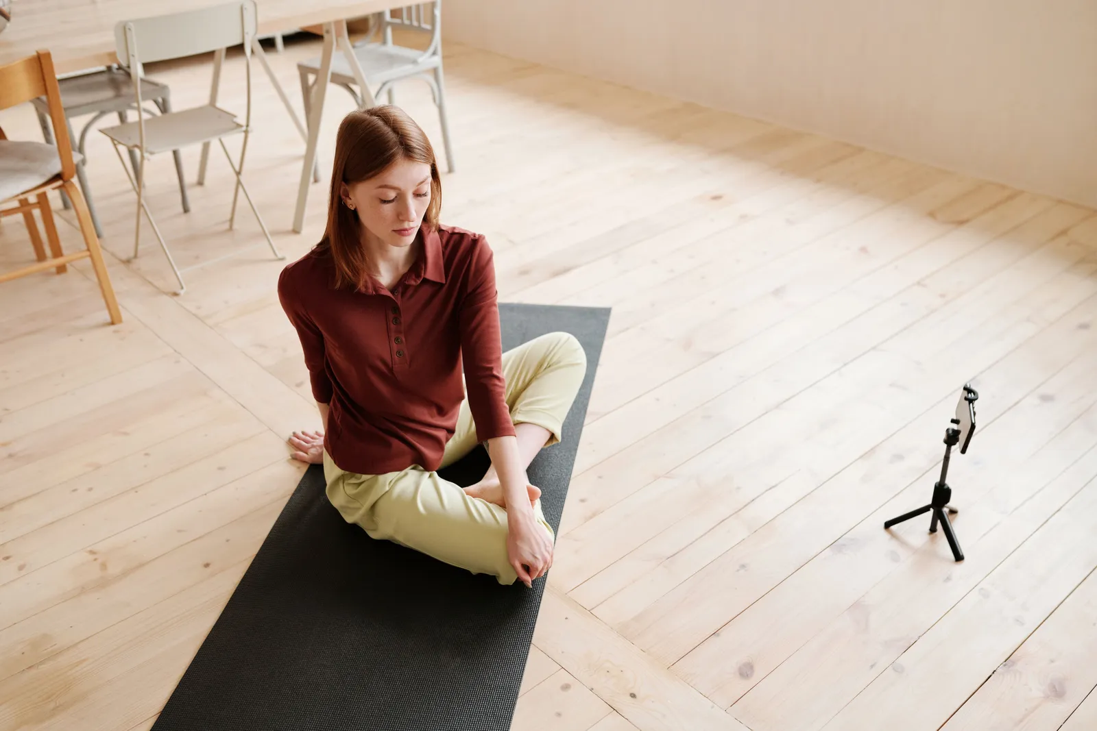 Young woman practicing yoga indoors with smartphone setup for recording, demonstrating flexibility and mindfulness