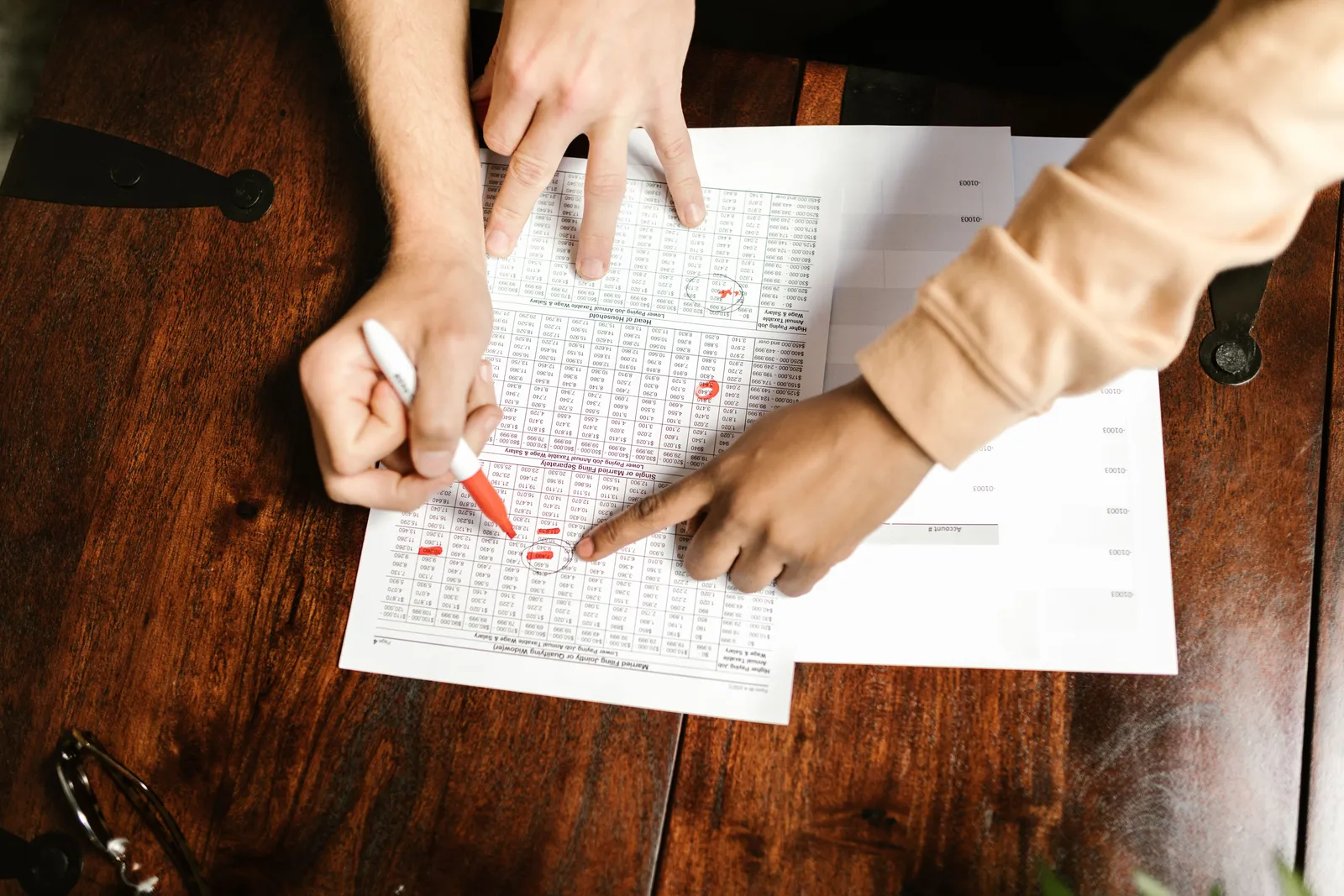 Two hands reviewing and marking financial documents with a red pen on a wooden table, top view