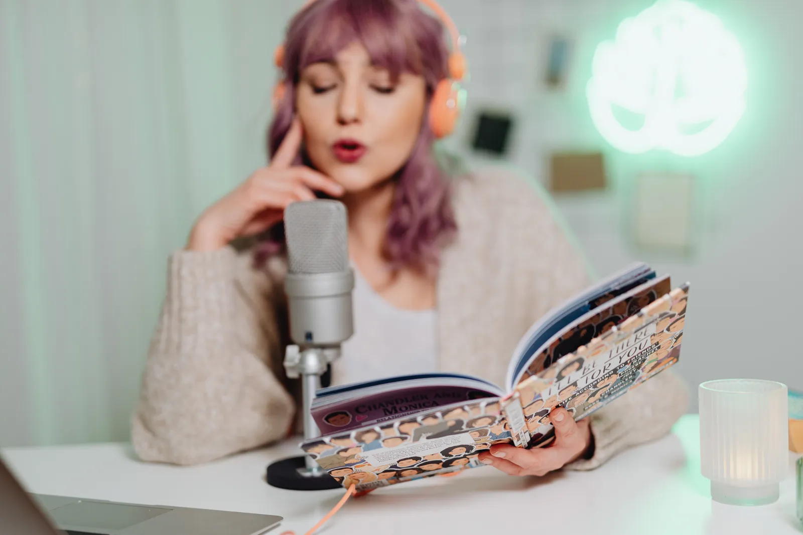 A woman wearing headphones reads a book into a microphone, recording a podcast indoors