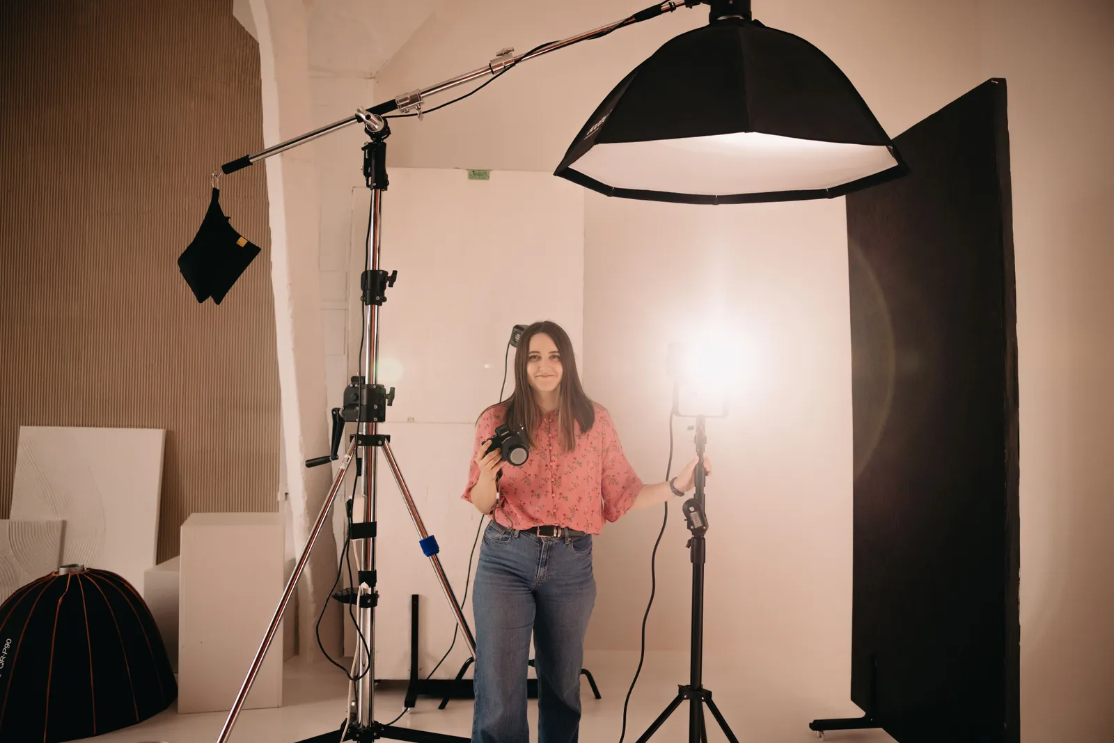 Female photographer holding camera in a well-lit studio with professional lighting and equipment