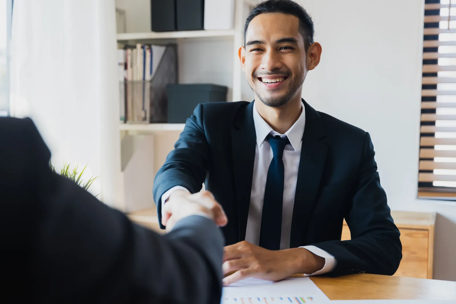 Confident businessman in suit shaking hands at office desk, symbolizing successful partnership
