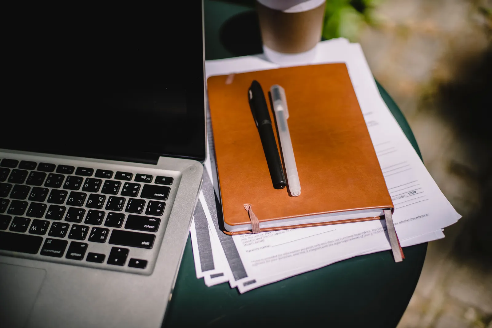 From above of leather notebook with papers and pens placed on table near laptop and cup of coffee