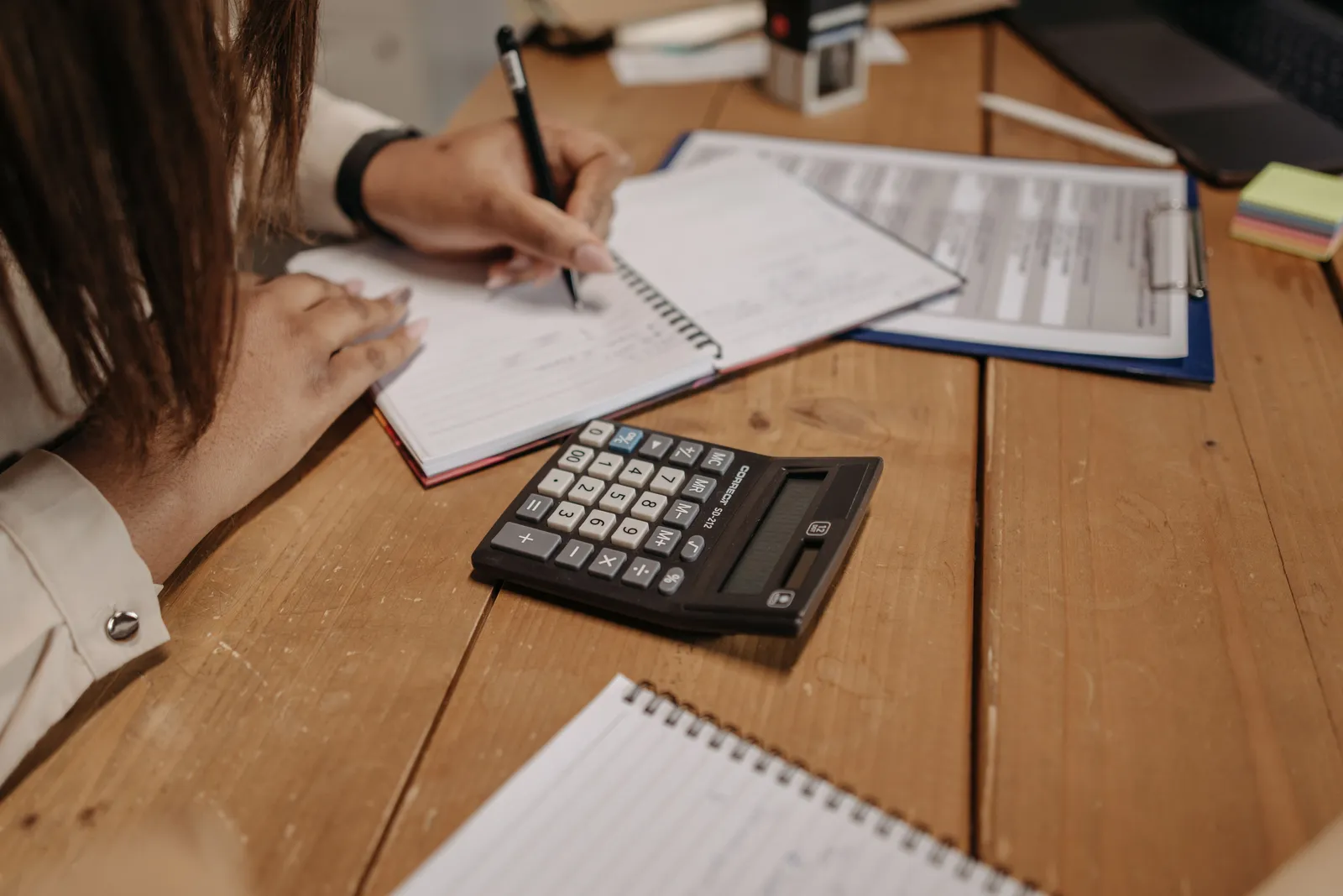 Woman writing notes with a calculator on a wooden desk, focusing on work and accounting