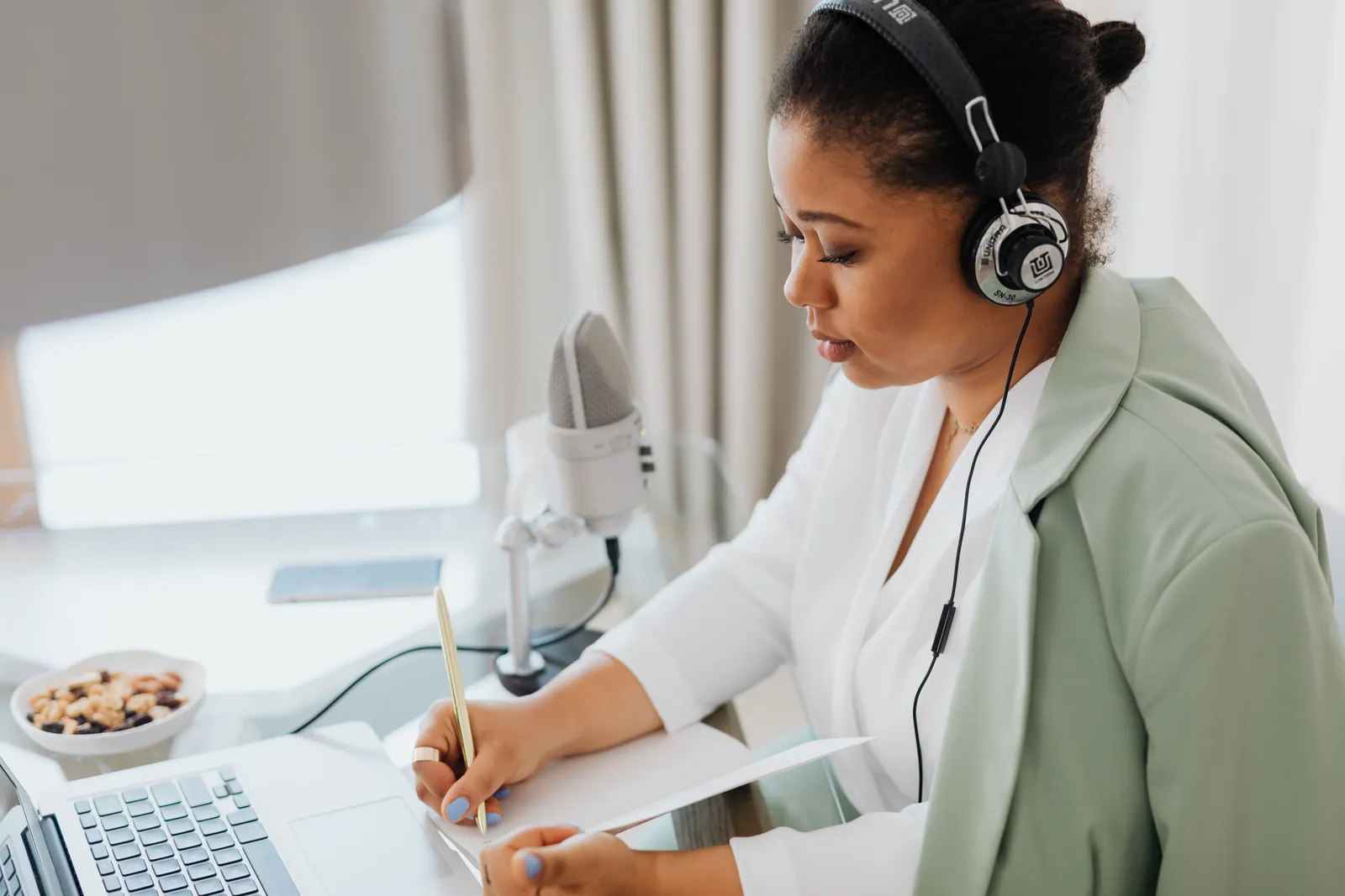 Woman wearing headphones, writing notes, and working on a laptop indoors