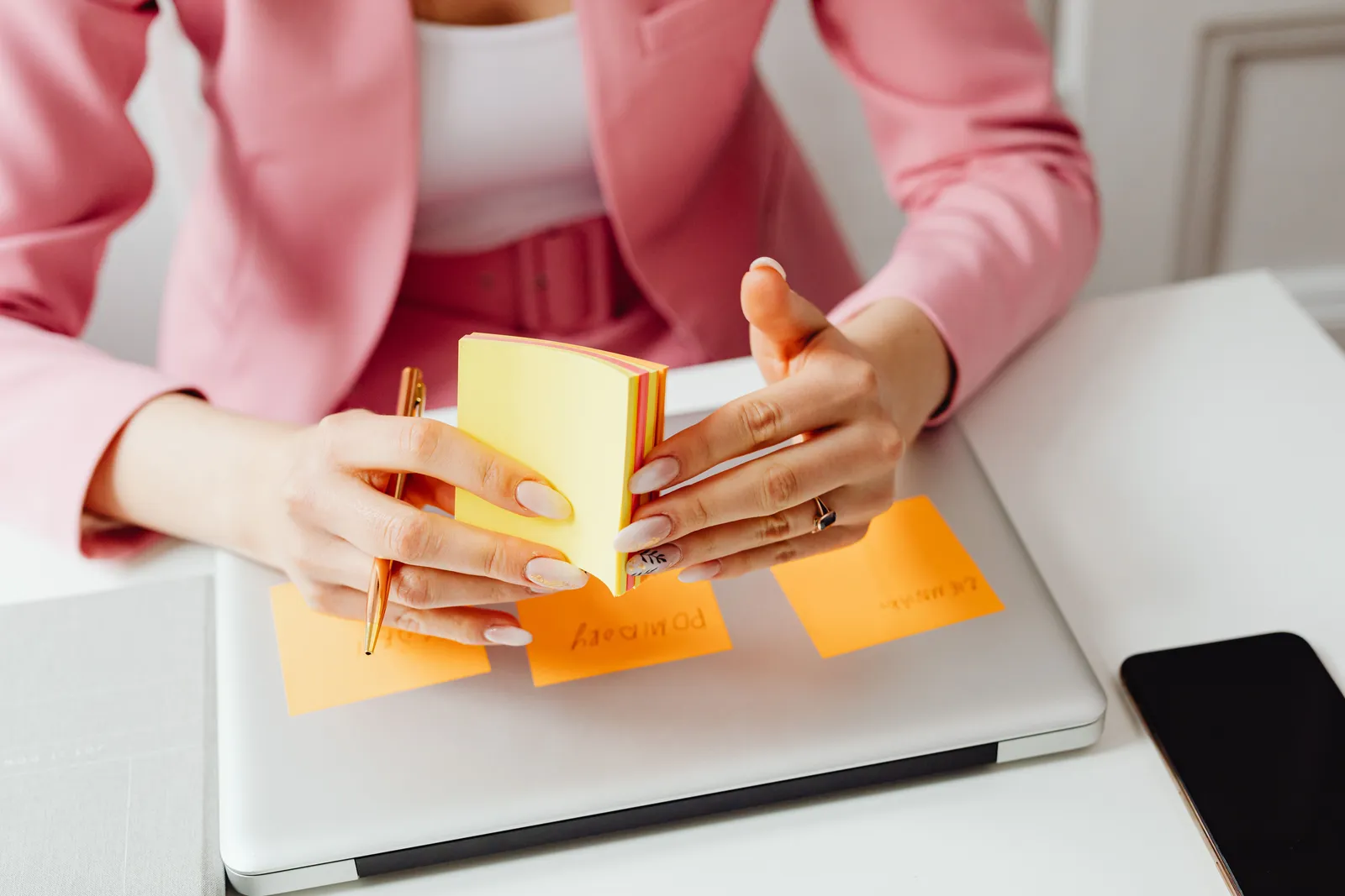 A woman in a pink blazer arranges sticky notes on her laptop, focusing on organization and planning