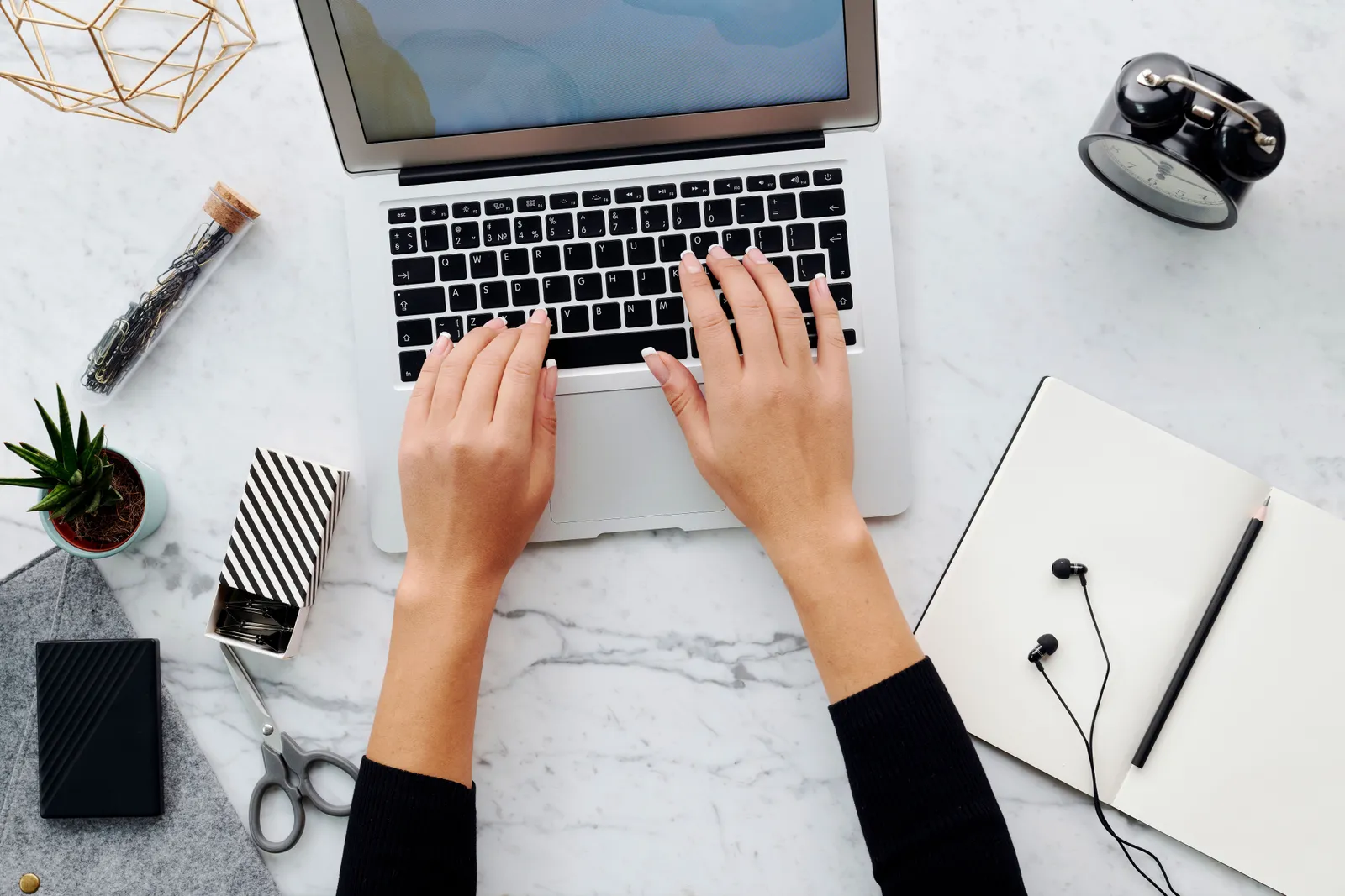 Hands typing on a laptop with various office supplies on a marble desk