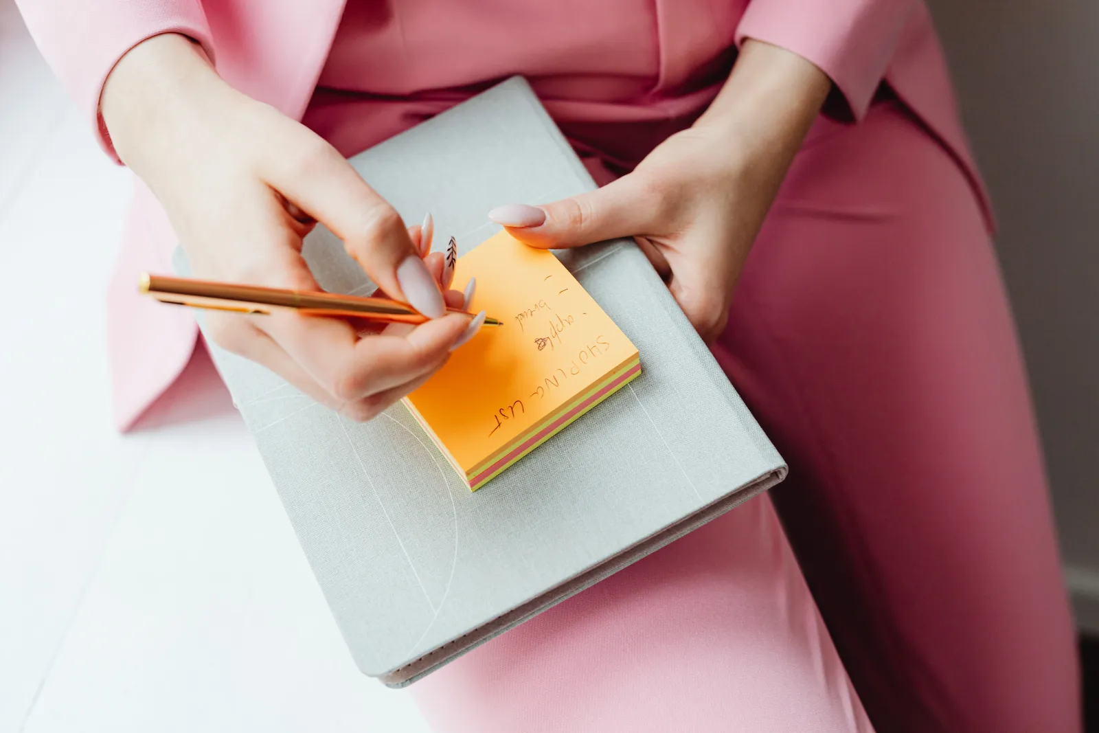 Close-up of a woman writing on a sticky note with a pencil, wearing a pink suit