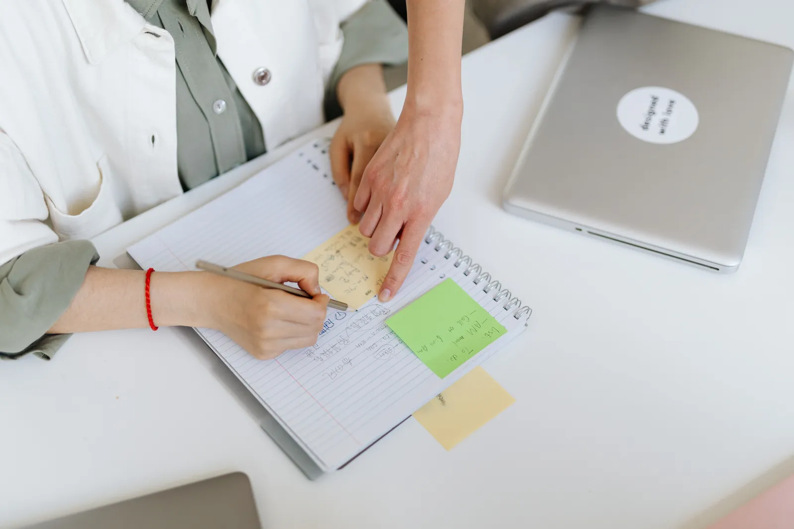 Two people collaborating over a notebook with post-it notes and a laptop in an office setting