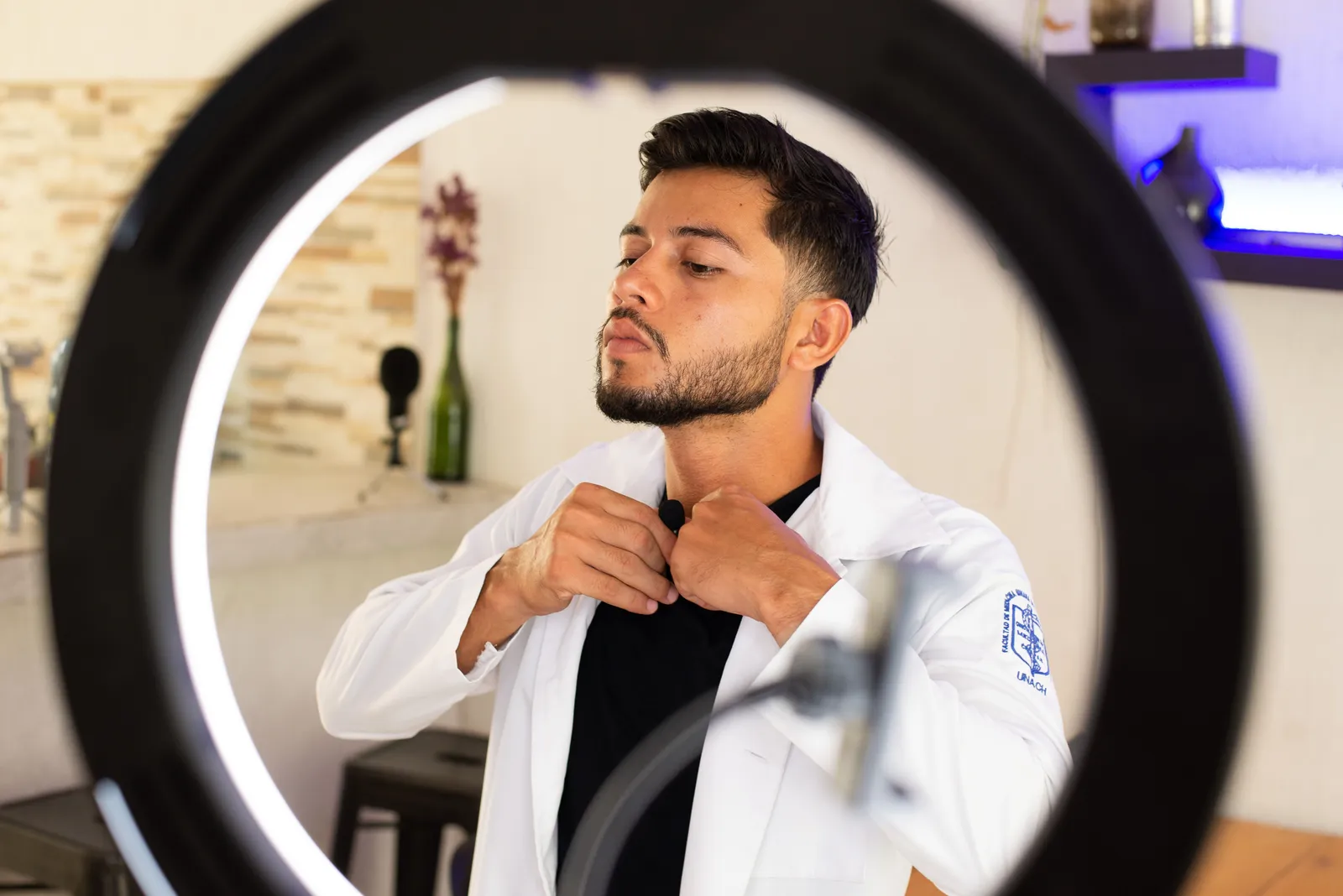 A young man adjusting his lapel mic in a studio with a ring light, preparing for a video shoot