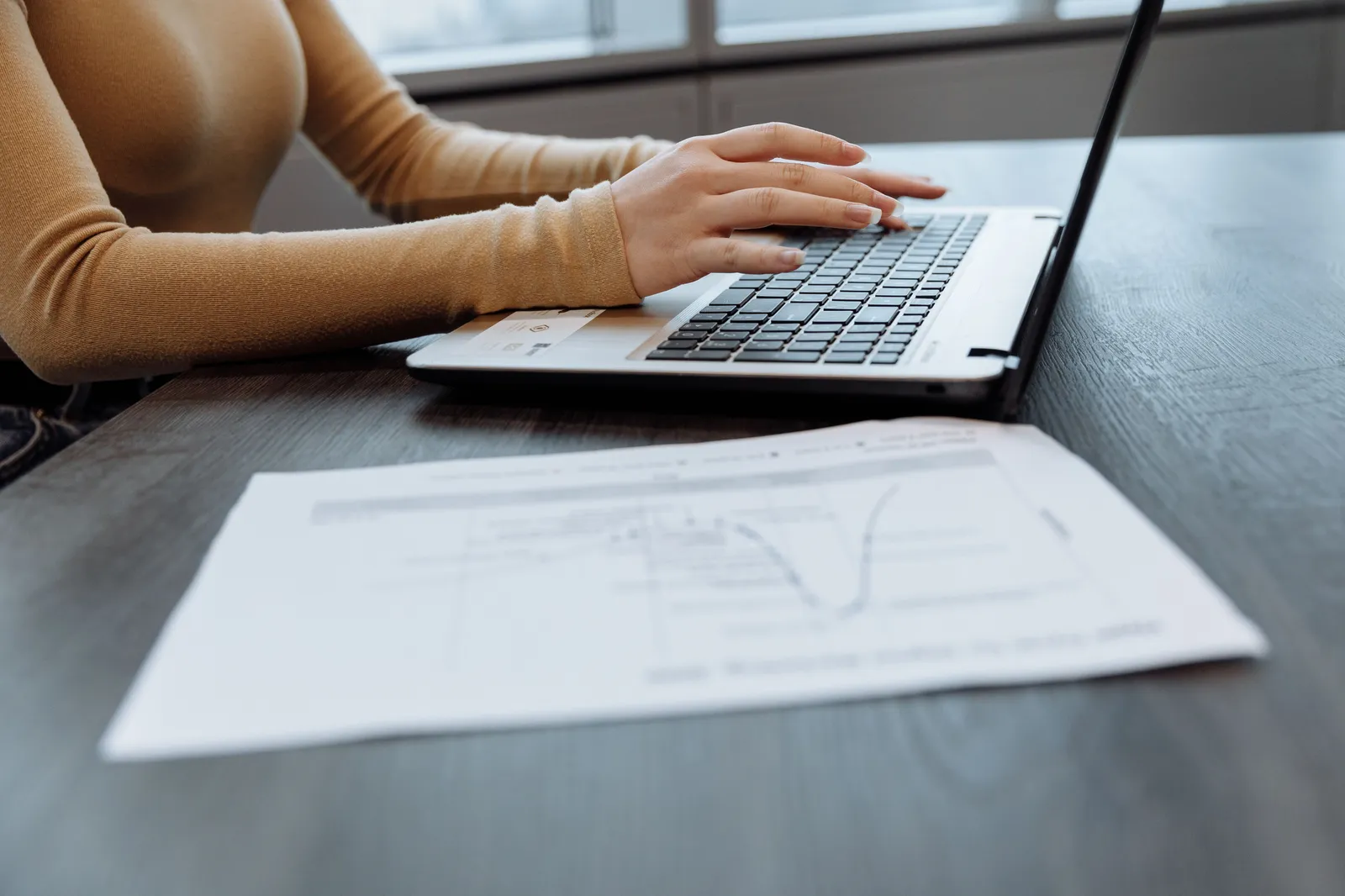 Close-up of a person's hands typing on a laptop at a desk with documents. Ideal for office, work, and technology themes