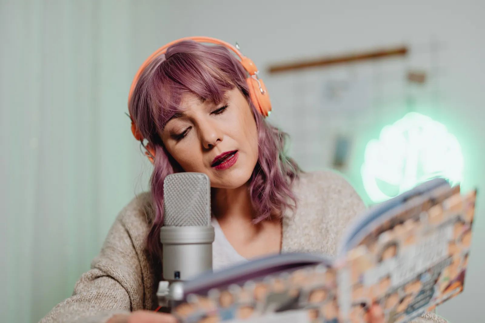 A woman wearing headphones reads from a book into a microphone indoors