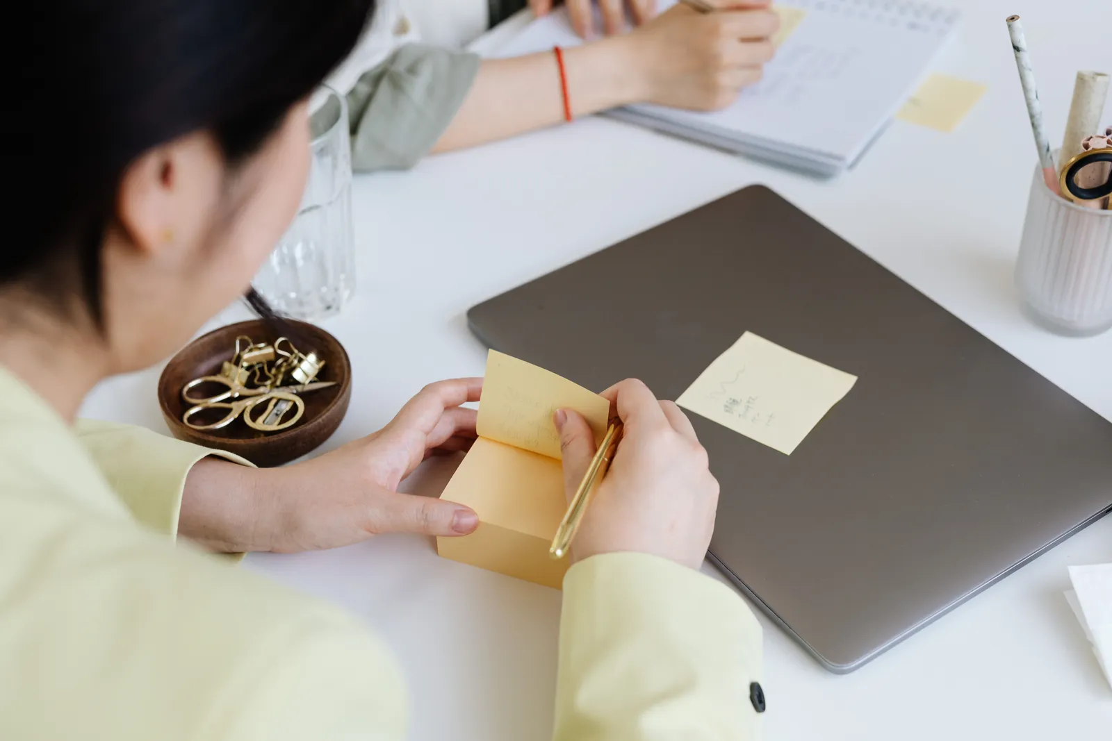 Close-up of a woman writing notes at a desk with a laptop and stationery items