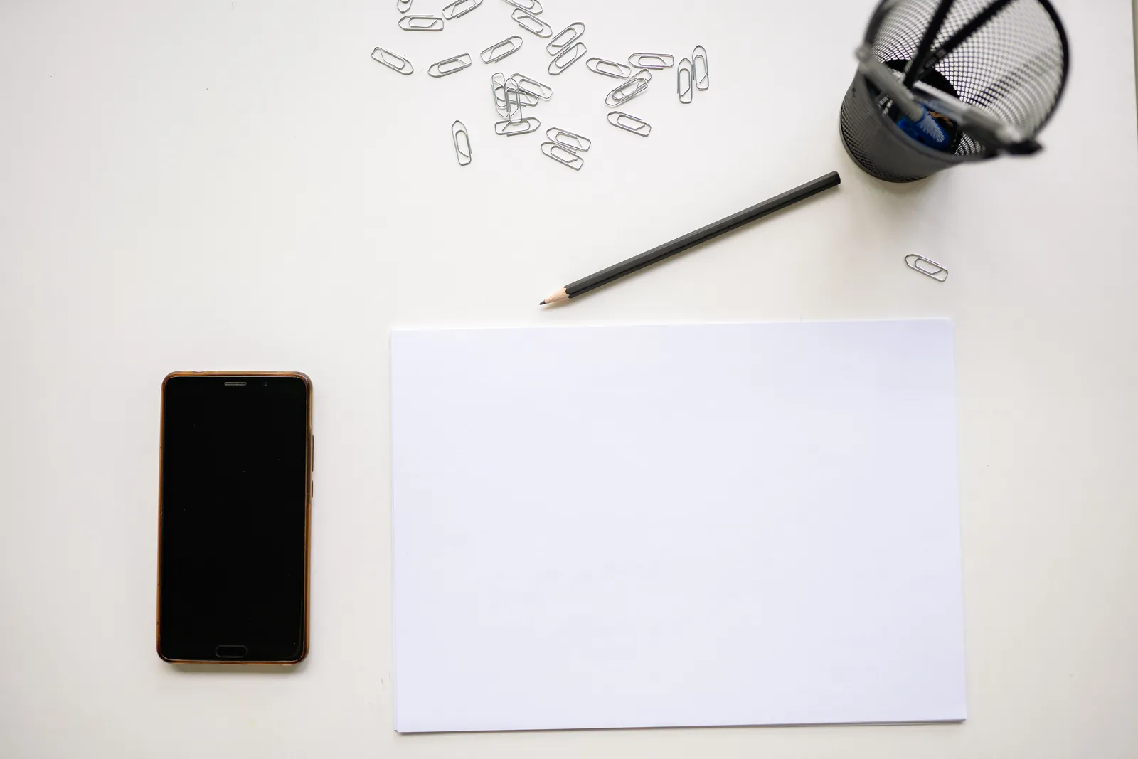 Top view of a minimalist desk layout with smartphone, pencil, paper clips, and stationery essentials on white paper