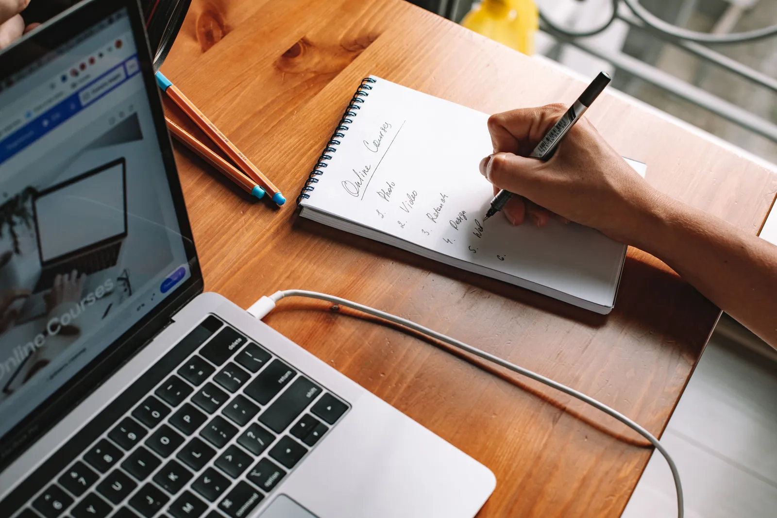 A person writing notes while researching online courses on a laptop in a home setting