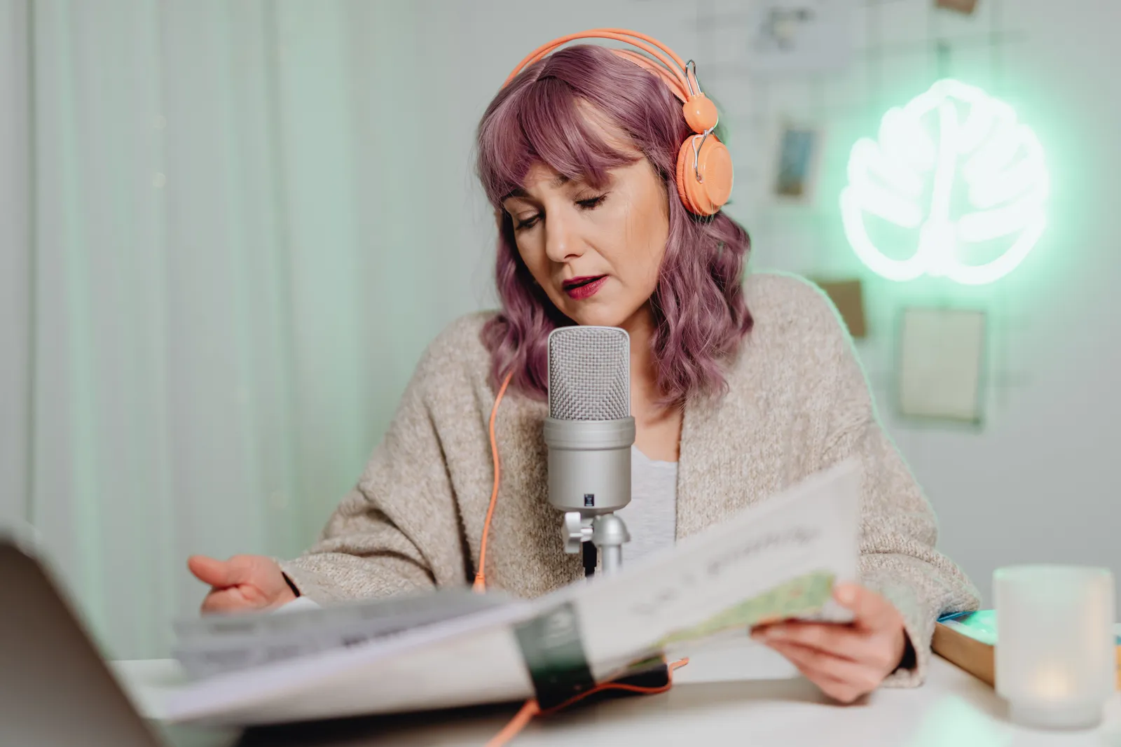 Woman with headphones podcasting indoors, speaking into a microphone while reading a book