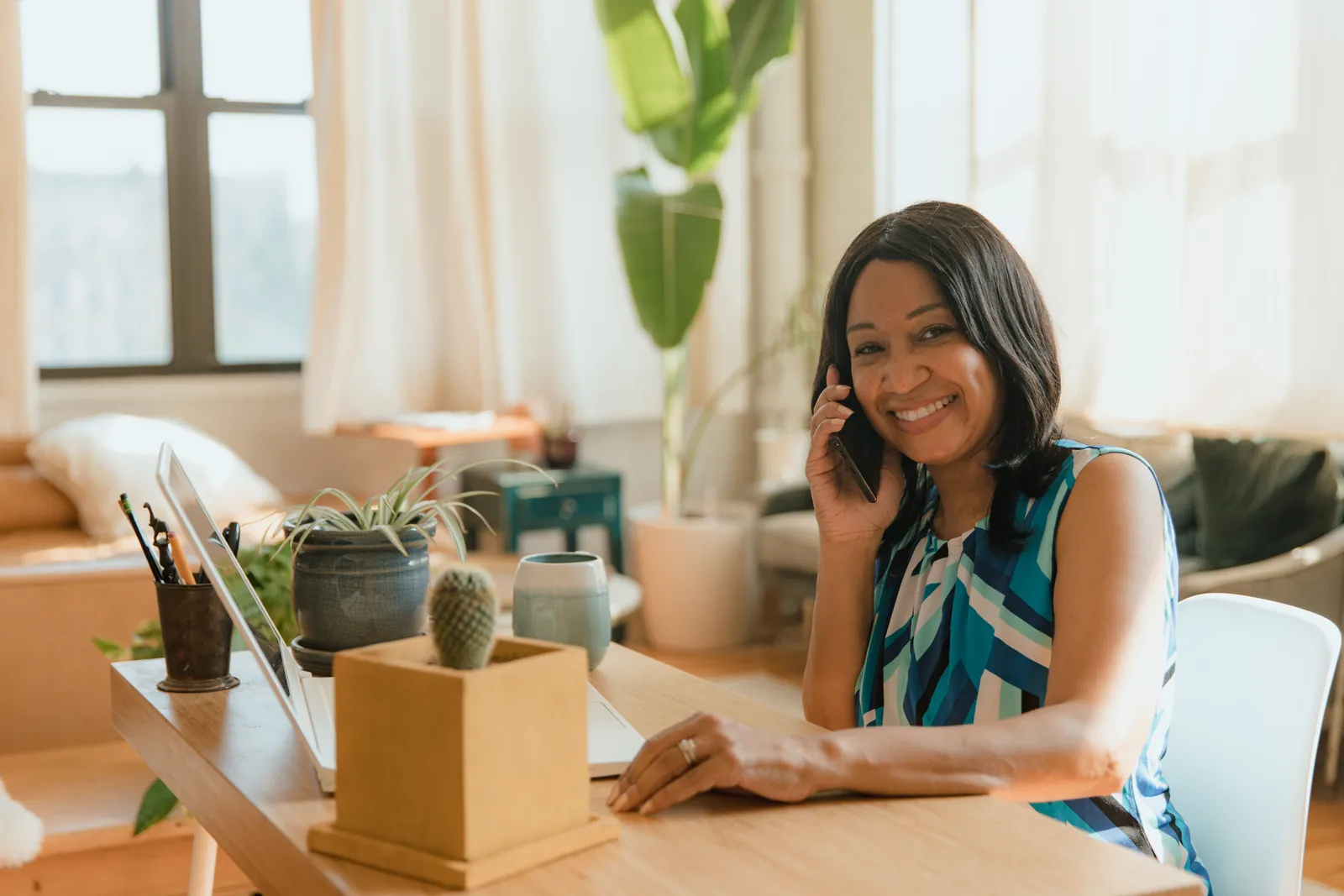 Smiling woman in home office talking on phone with laptop and plants on desk