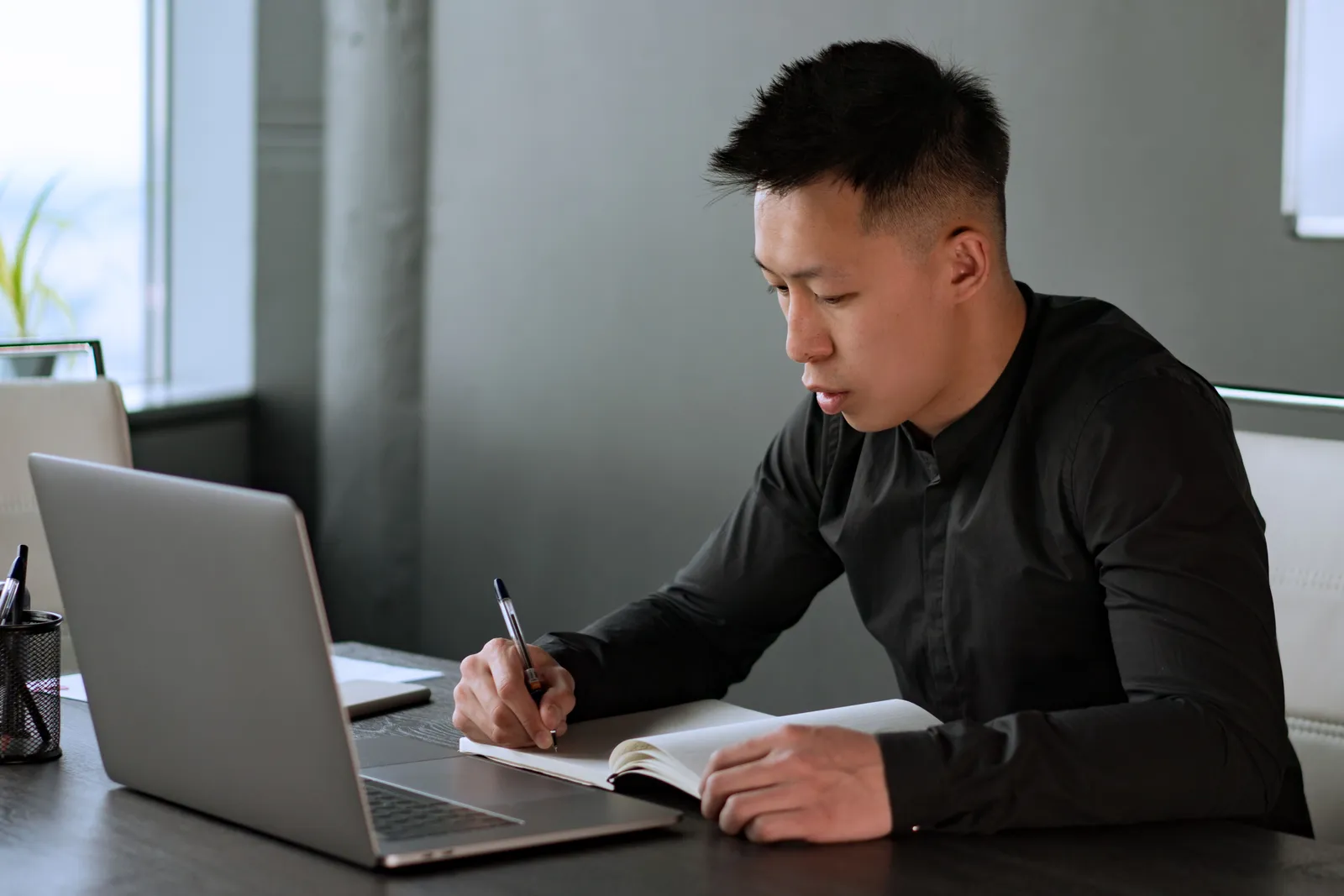 A professional man writes notes in a notebook at his desk with a laptop, demonstrating work focus