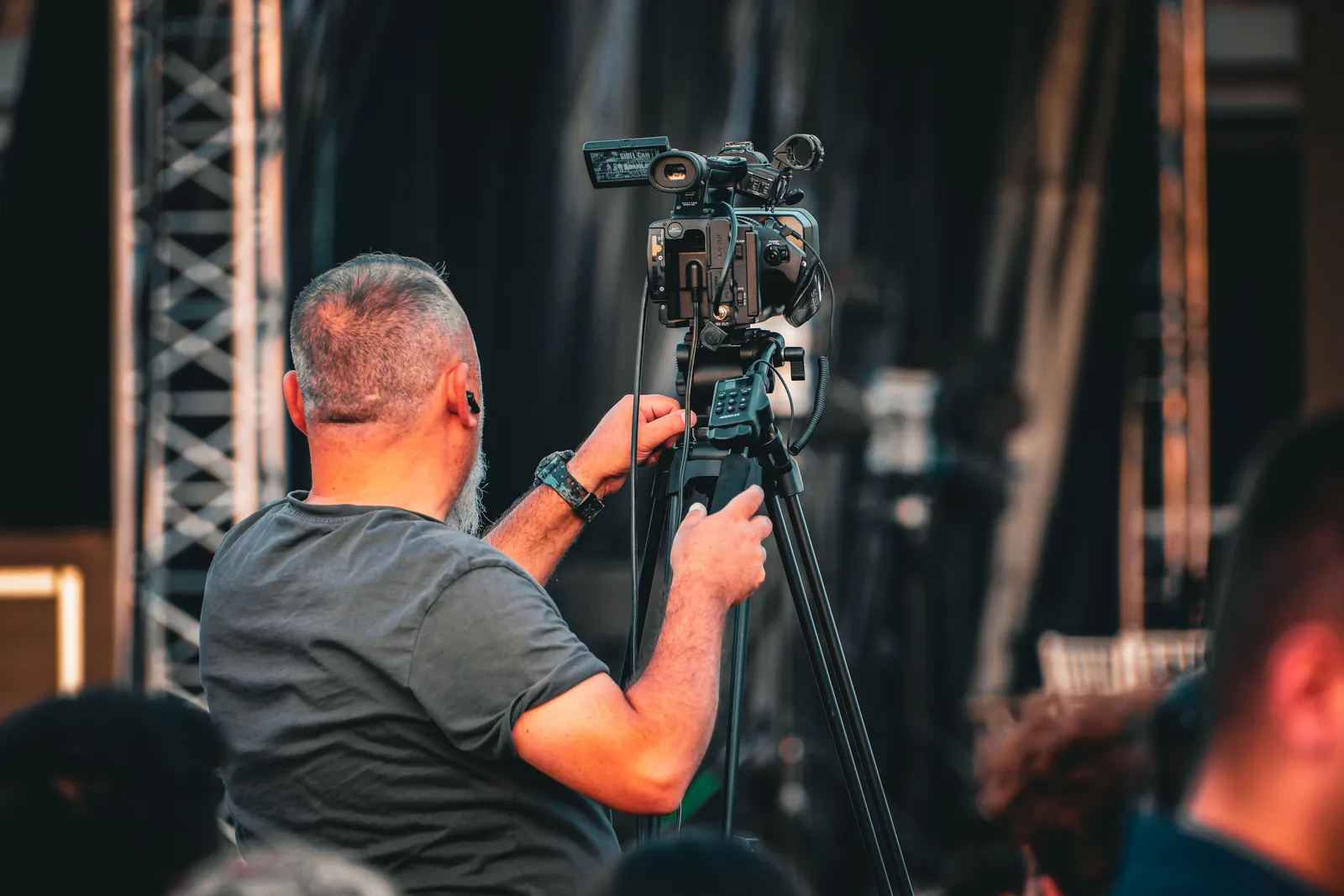 A professional cameraman operating a video camera on a tripod during an outdoor event, captured from behind
