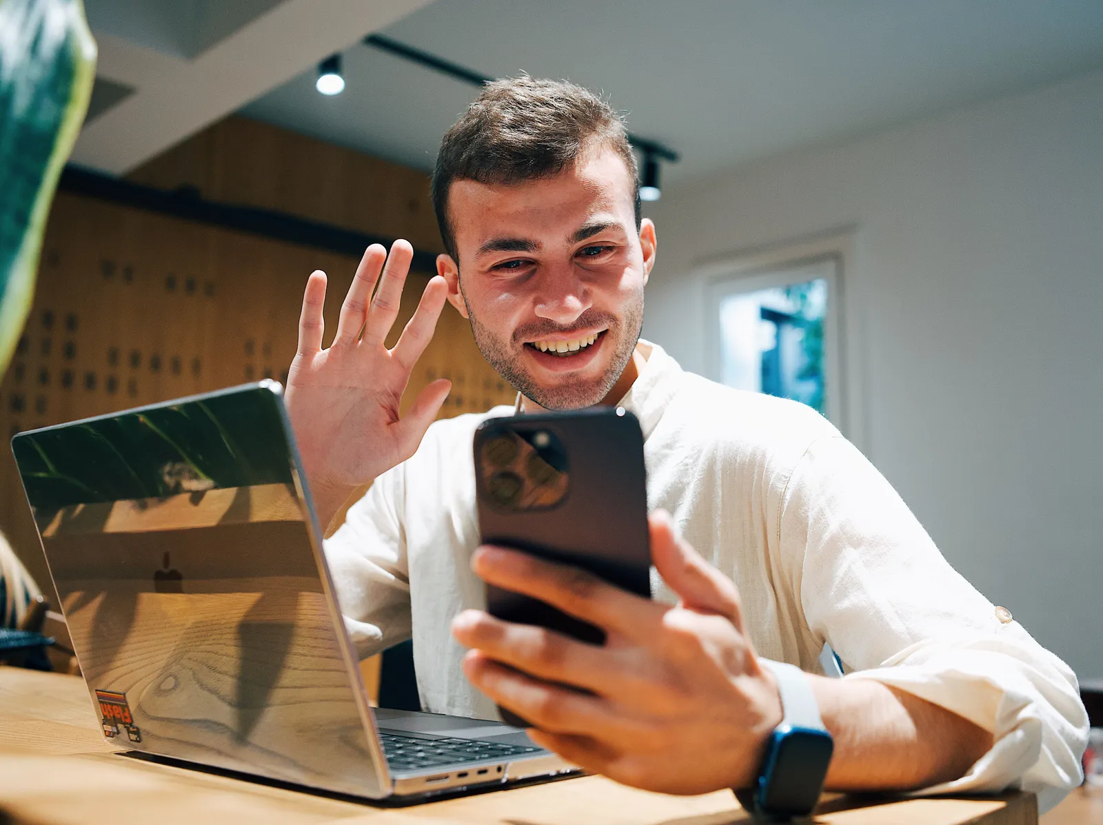 Smiling young man making a video call on his smartphone while sitting with a laptop in a modern office
