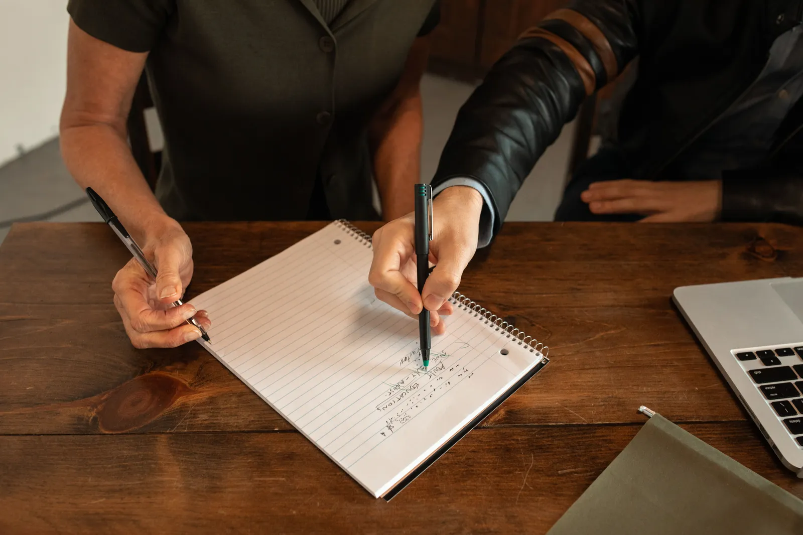 Two people writing notes during a collaborative meeting. Hands, notebook, and pens on wooden table