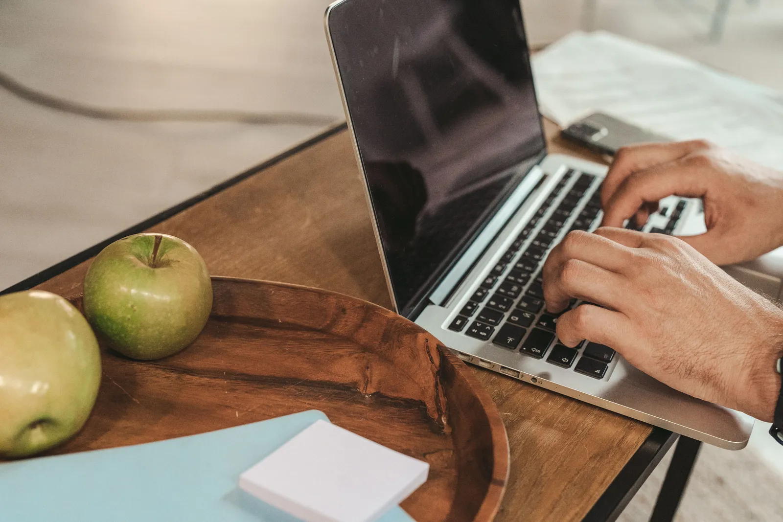 Hands typing on a laptop with green apples and sticky notes on a wooden desk, creating a productive work environment