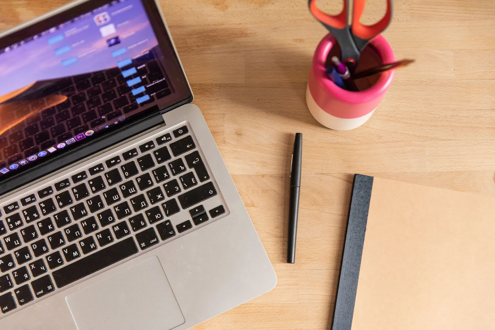 Top-down view of a laptop, pen, notebook, and scissors on a wooden desk, depicting a workspace setup