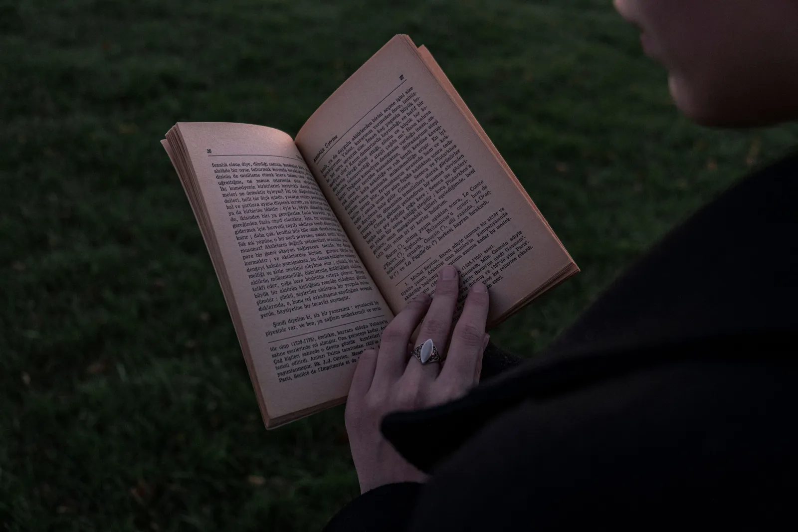Person holding and reading a book outdoors during twilight, focus on hands and open pages