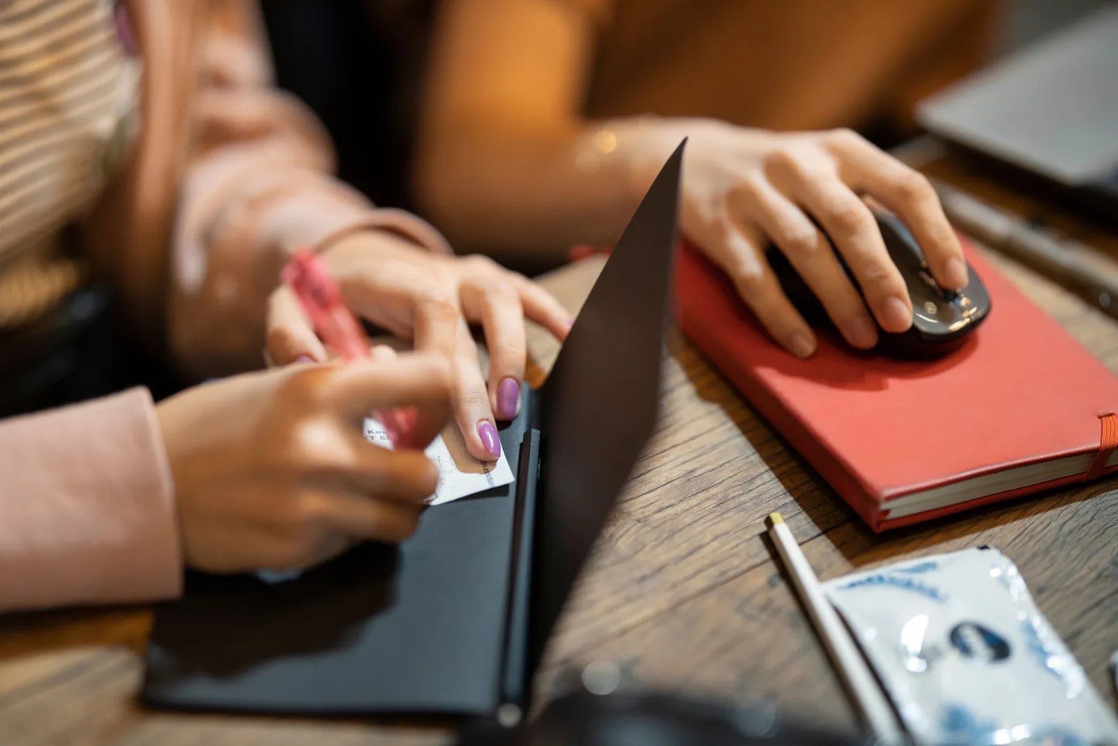 Close-up of hands writing in a notebook and using a computer mouse on a desk