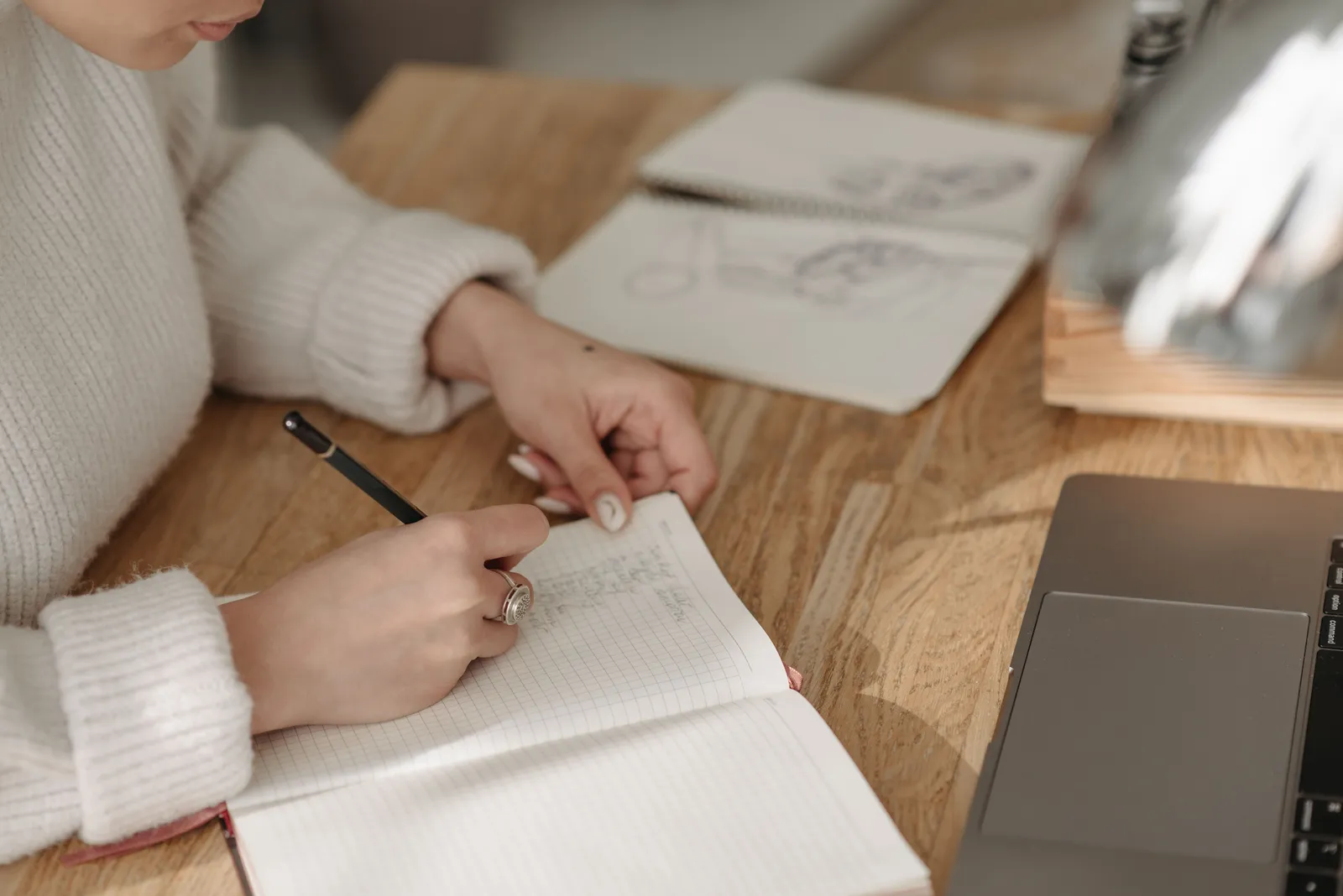A woman writes in a lined notebook at a wooden desk with a laptop and sketchbook nearby