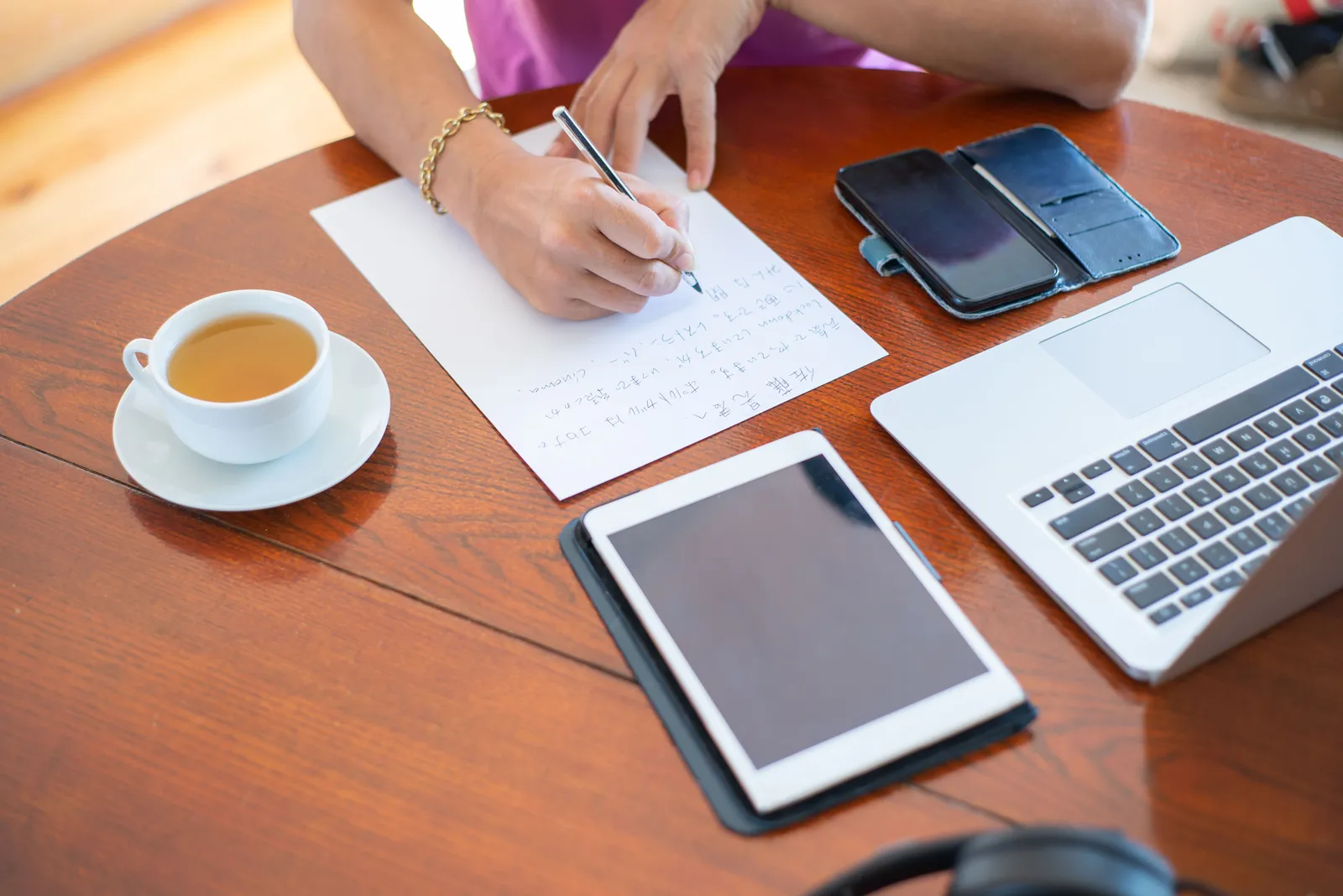 Close-up of a workspace with a laptop, tablet, smartphone, cup of tea, and a person writing notes
