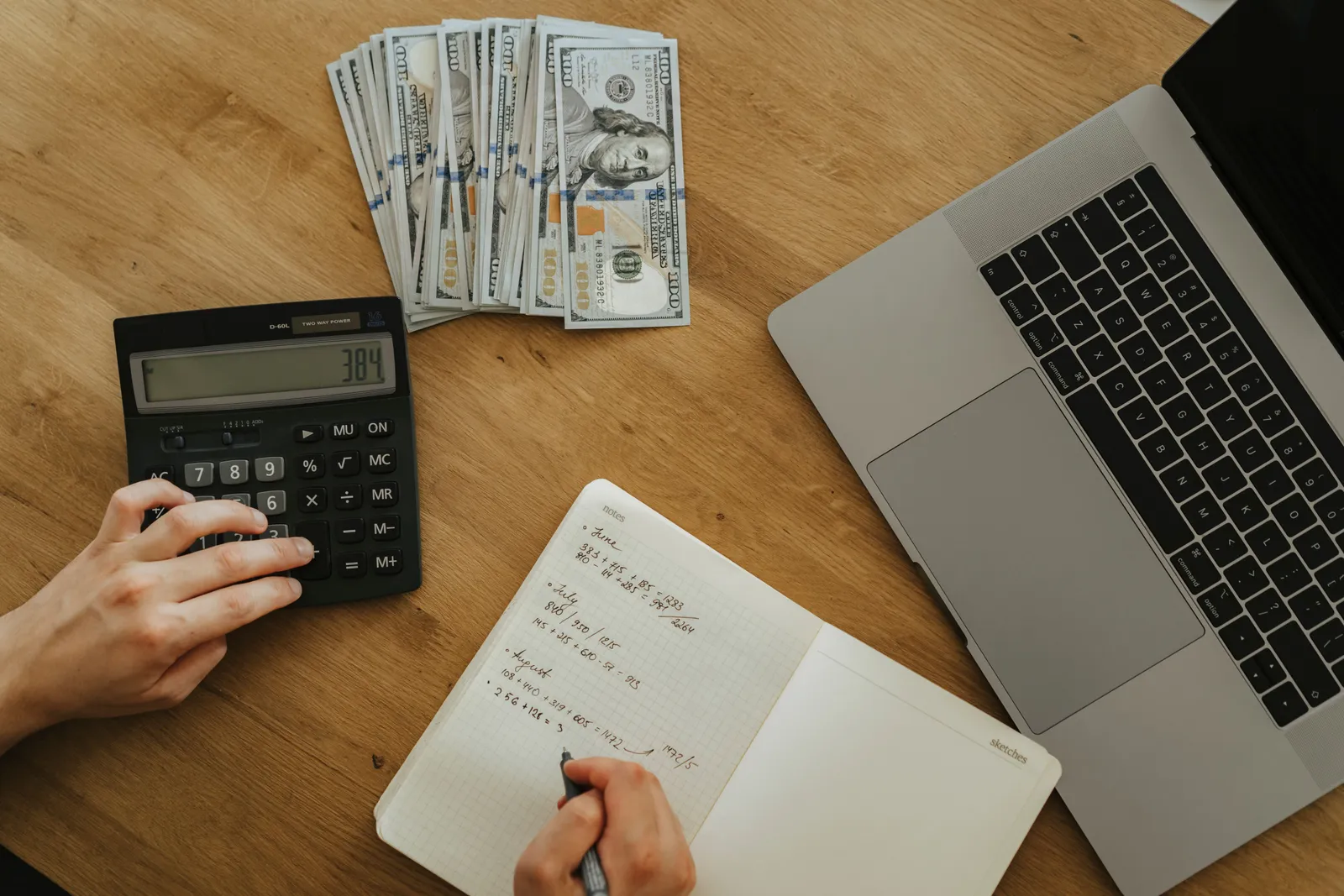 Hands using calculator and notebook, planning finances, with cash and laptop on wooden table