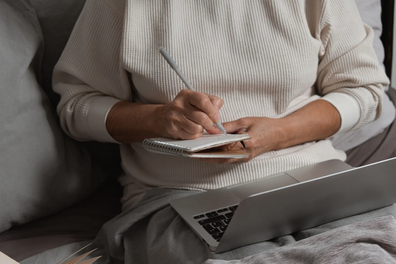 A person writing in a notebook with a pen while sitting by a laptop, creating a cozy work atmosphere