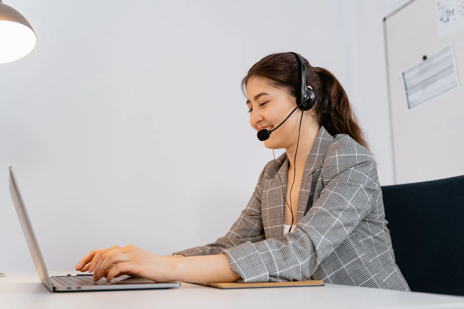 Woman in a plaid blazer working at a laptop with a headset in an office setting