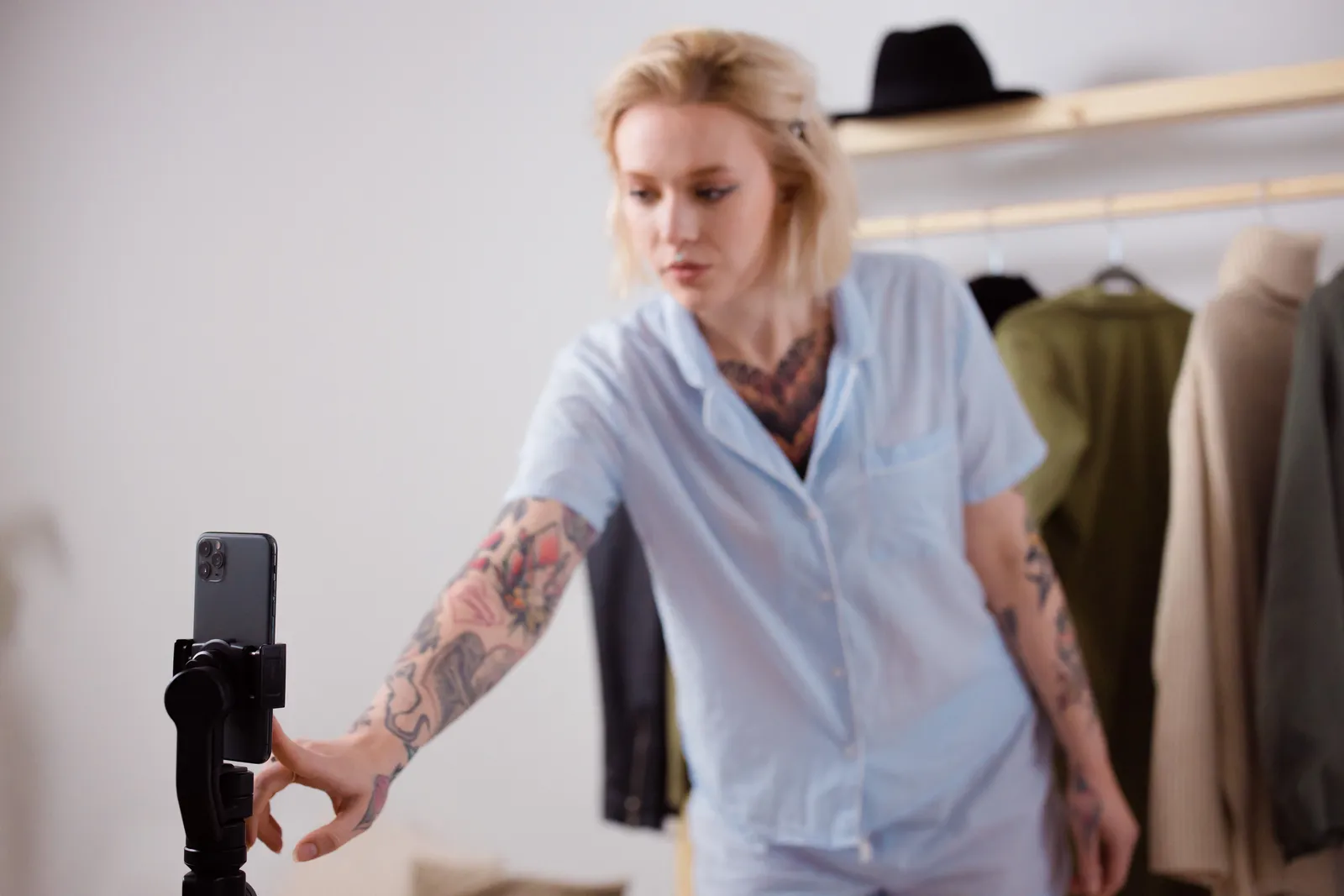 A stylish woman with tattoos adjusting her smartphone on a tripod, surrounded by clothing