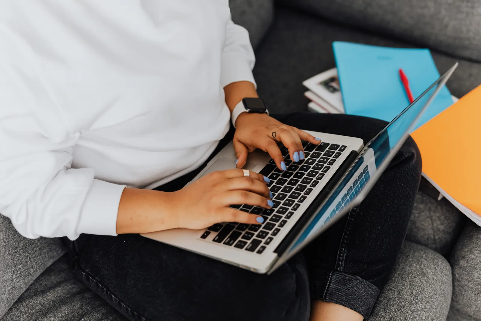 Person using a laptop on a gray sofa, surrounded by colorful notebooks, concept of remote work or study