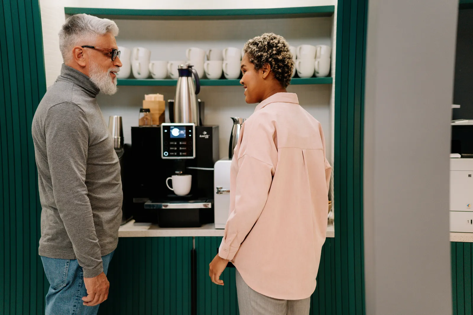 Senior and adult colleagues enjoying a coffee break, chatting by the coffee machine in an office break room