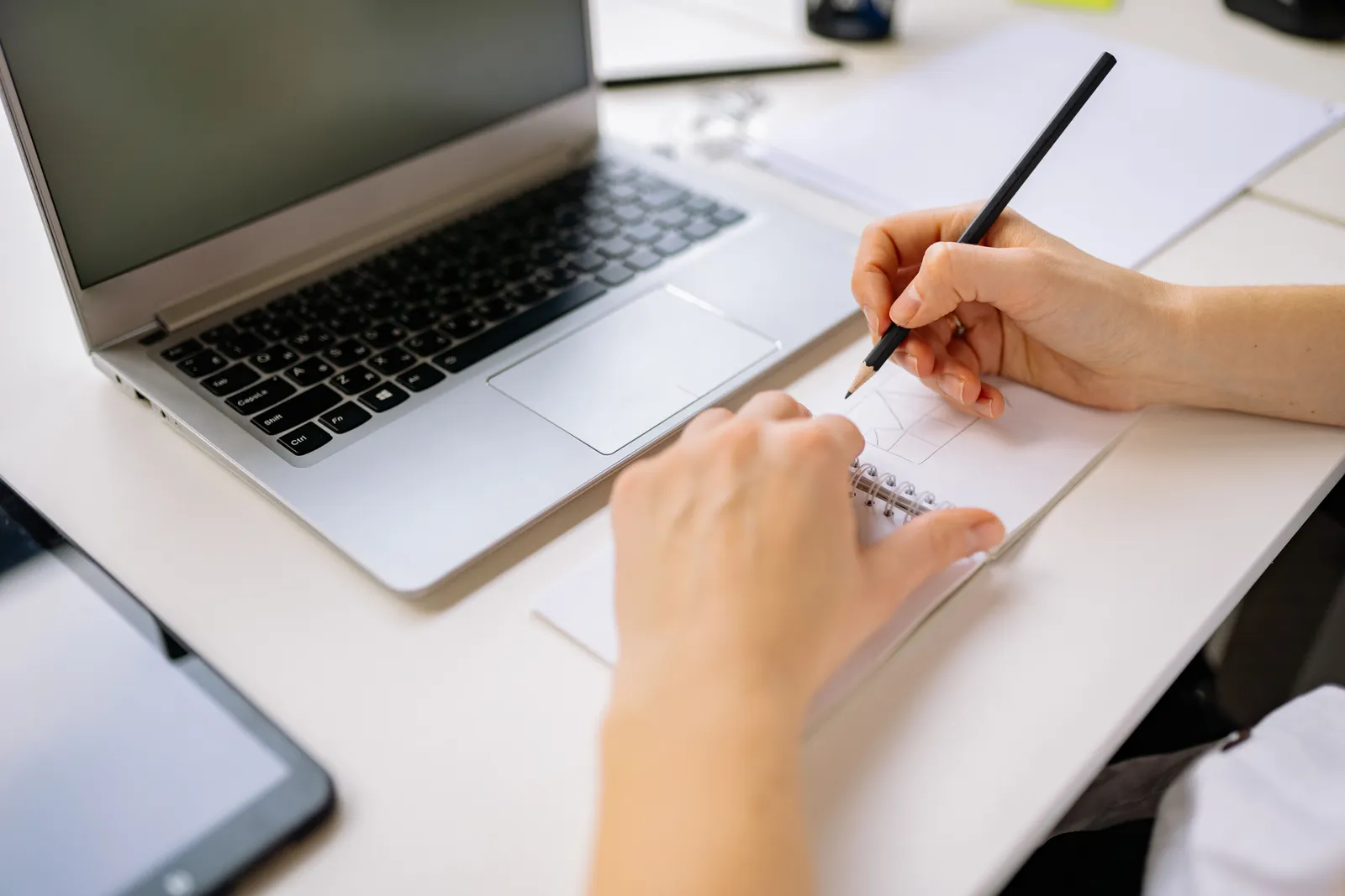 Hands sketching in a notebook beside a laptop on a desk, capturing creativity