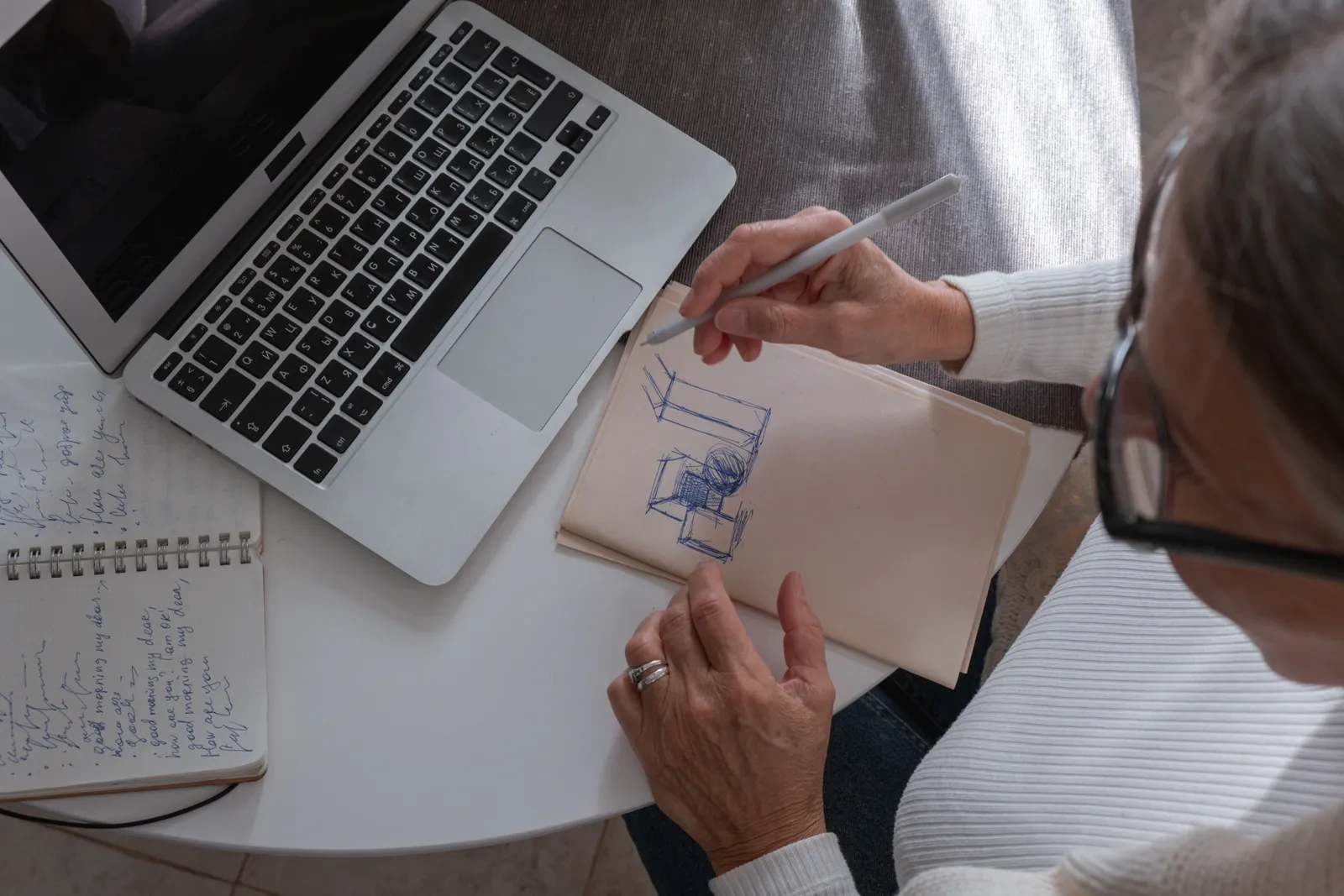 An adult woman sketching in a notebook at a desk with her laptop, working from home